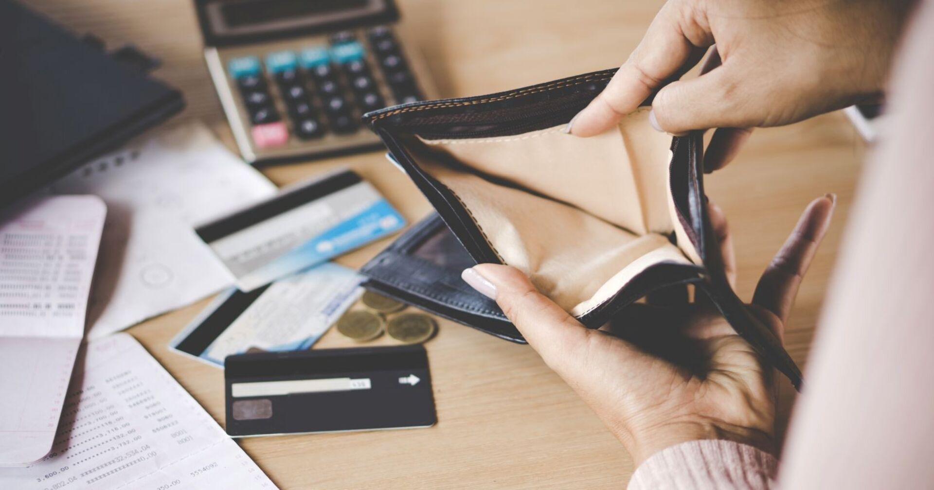Frau öffnet leere Geldbörse über einem Holztisch, auf welchem Geld Kreditkarten und Rechnungen liegen