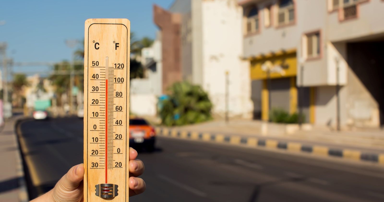 Hand hält Thermometer, das über 40 Grad Celsius zeigt, vor einer sonnigen Straße mit Gebäuden und Autos.