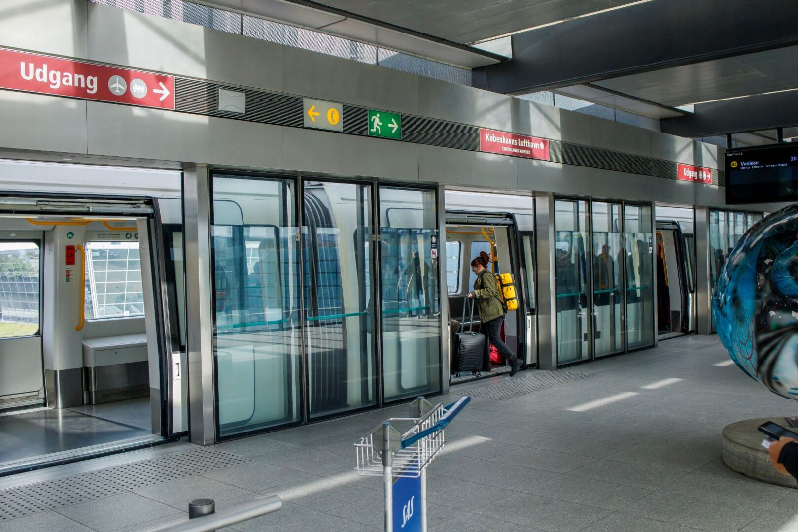 Fahrerlose U-Bahn der Kopenhagener Metro an der Station „Københavns Lufthavn“ mit einsteigender Passagierin und modernen Bahnsteigtüren.