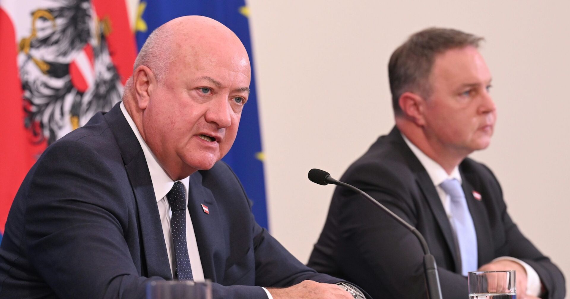 Christian Stocker und Andreas Babler sitzen bei einer Pressekonferenz vor Mikrofonen. Im Hintergrund sind die Flaggen Österreichs und der EU zu sehen.