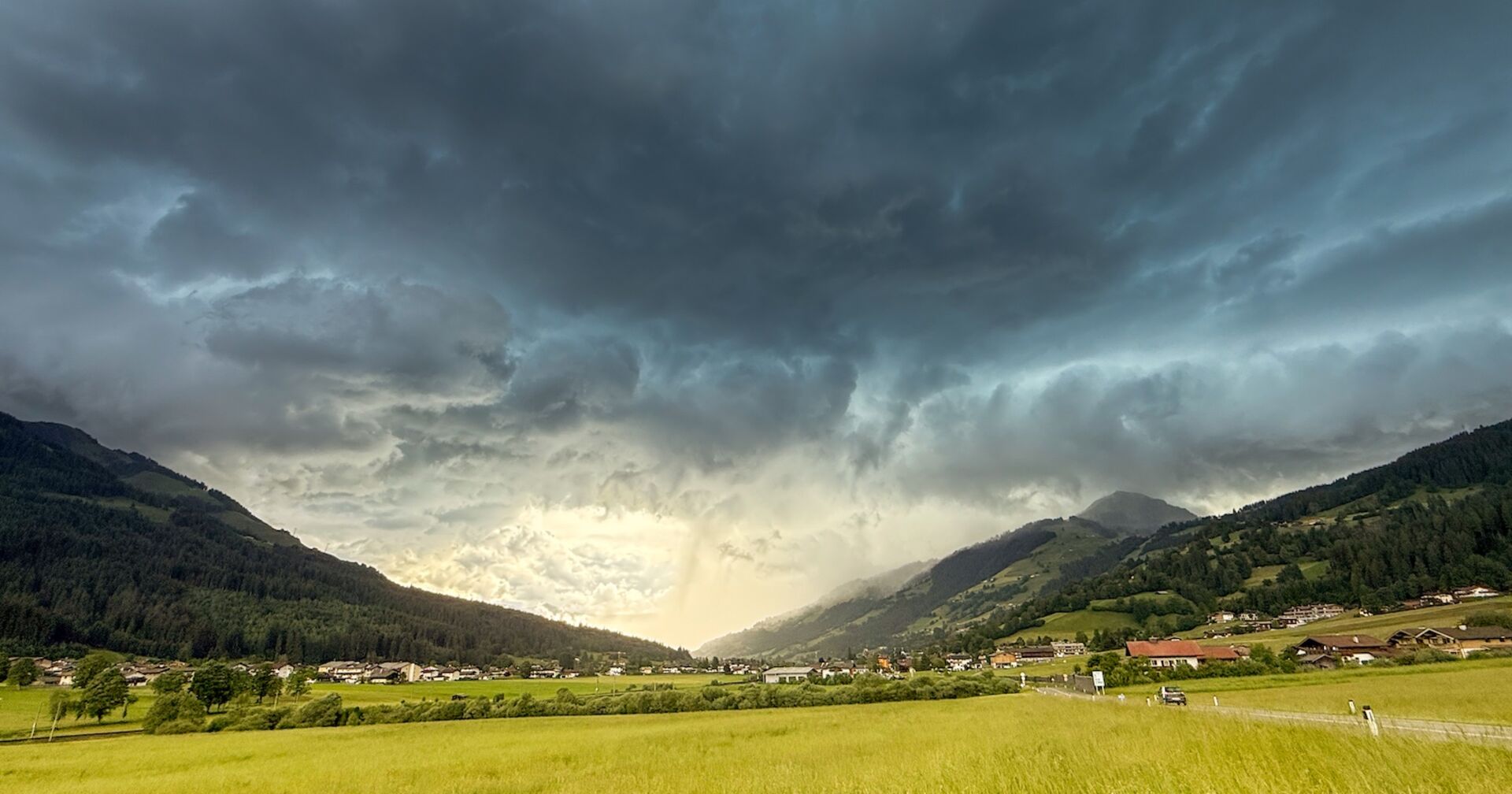 Dunkle Gewitterwolken über einem alpinen Tal, im Vordergrund grüne Wiesen und ein kleines Dorf; Unwetterfront im Anmarsch.: Katastrophale Unwetter zogen in den Abendstunden am Alpenrand entlang. Besonders heftig betroffen war der Landkreis Garmisch-Partenkirchen und Tirol. Am Achensee kurz vor der deutschen Grenze fiel Golfballhagel.