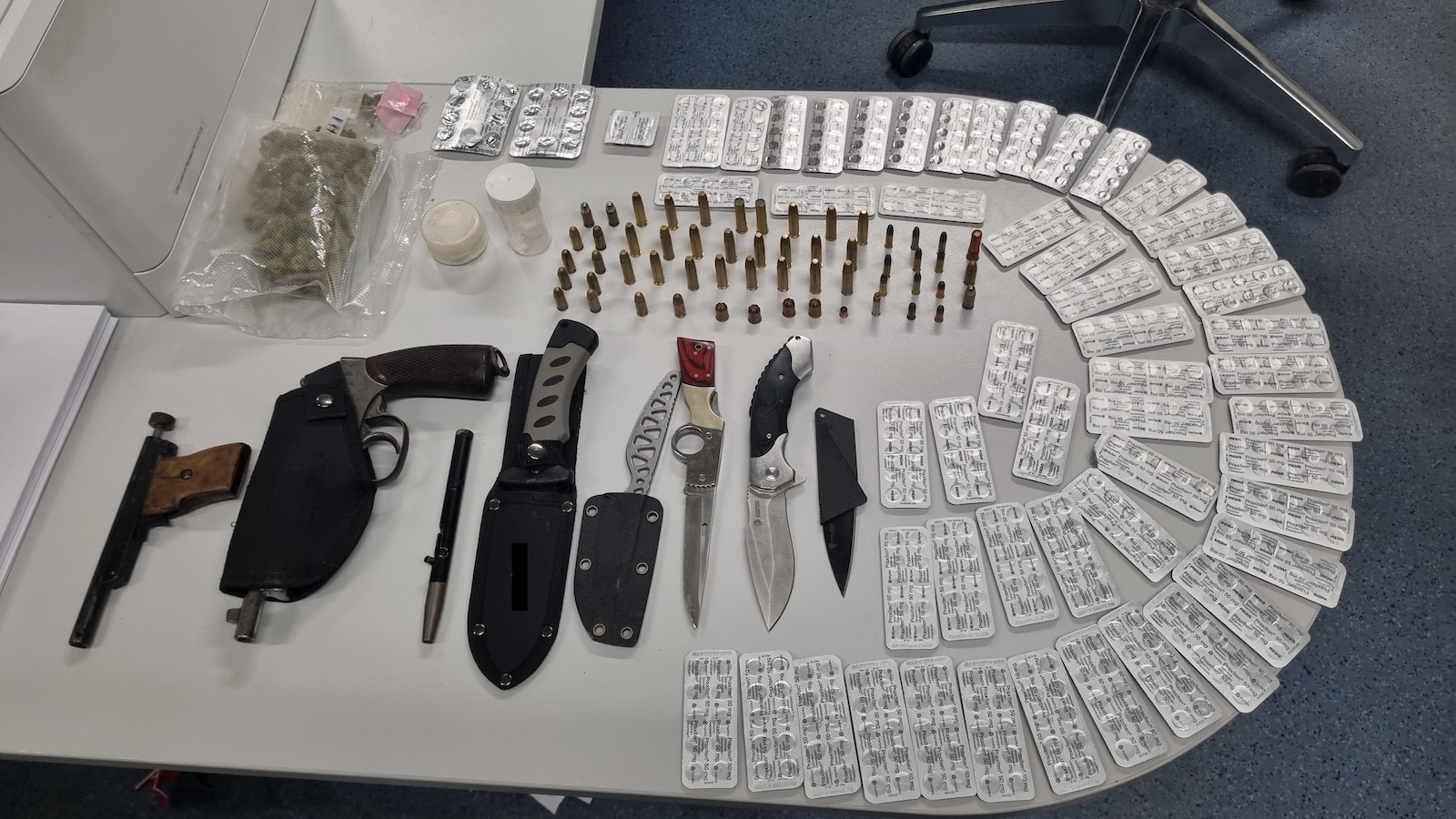 Sichergestellte Waffen, Messer, Munition, Tabletten und Drogen auf einem Tisch in einem Polizeibüro.