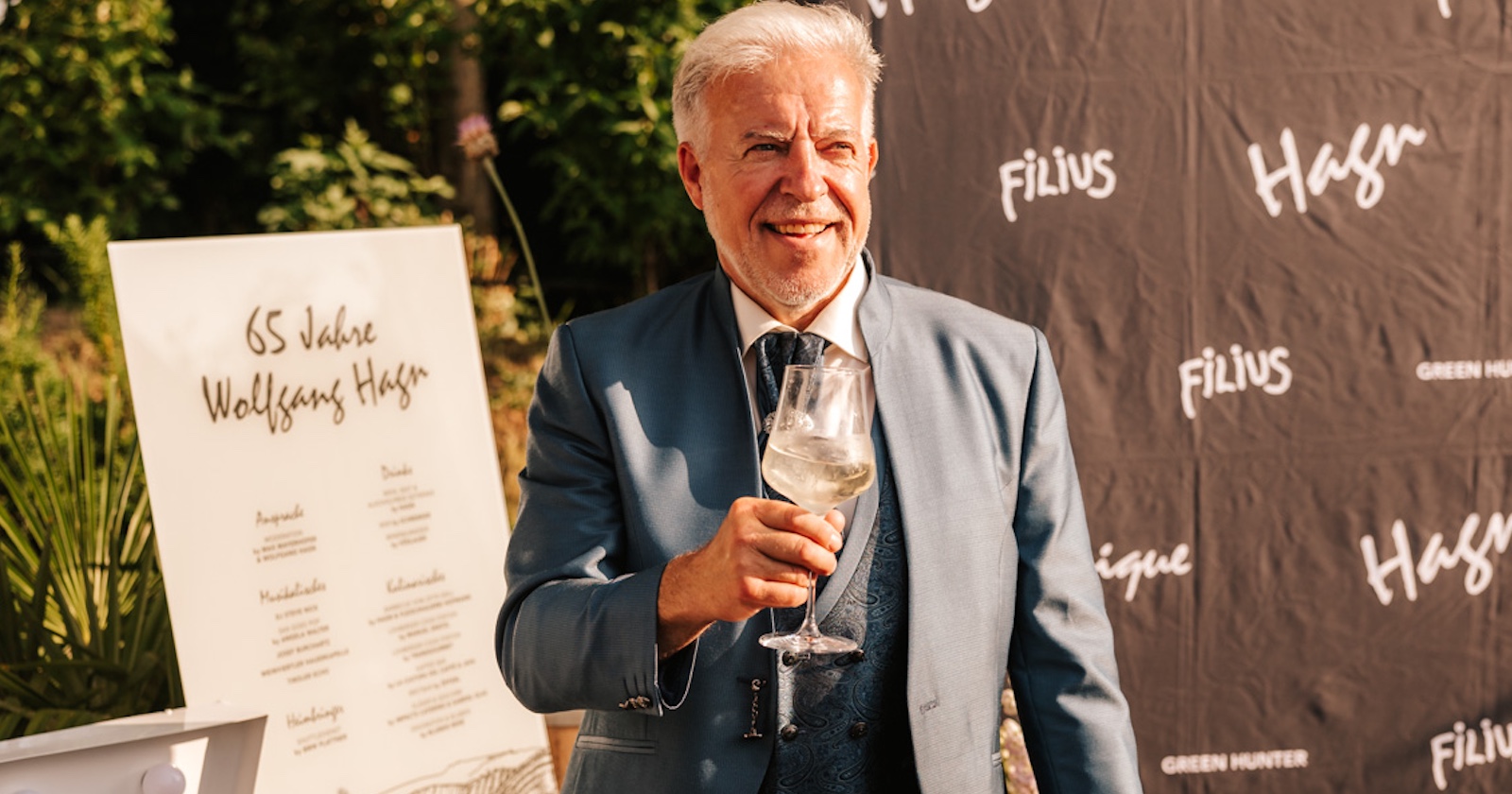 Mann mit weißen Haaren im grauen Festagsanzug und einem Glas Wein