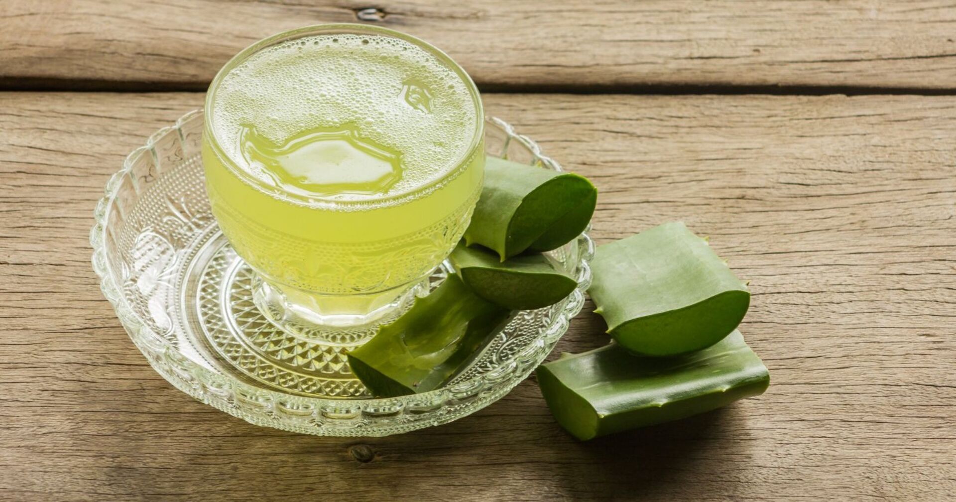 Ein Glasteller und ein Glas stehen auf einem Holztisch. Im Glas befindet sich eine hellgrüne Flüssigkeit. Am Teller und am Tisch liegen einige Stücke Aloe Vera.