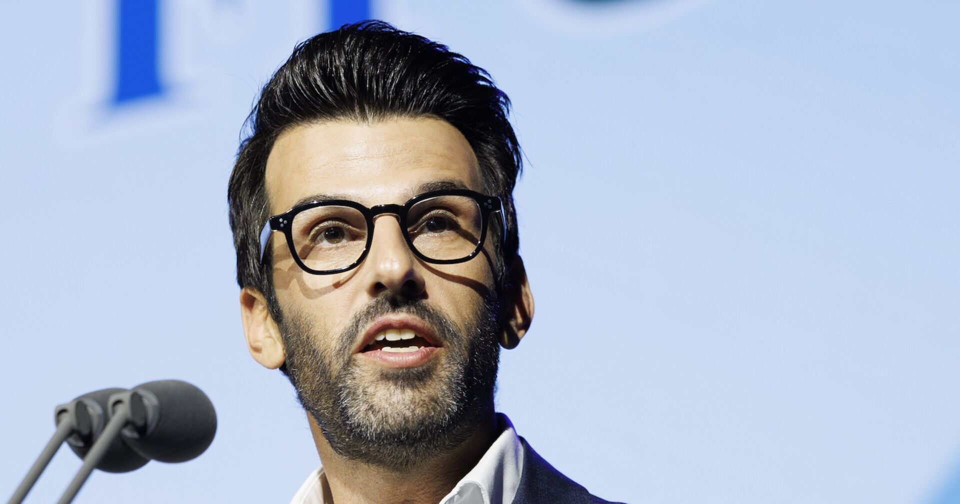 Udo Landbauer spricht vor Mikrofon bei Pressekonferenz, trägt Brille und Anzug, Hintergrund mit unscharfem FPÖ-Logo.