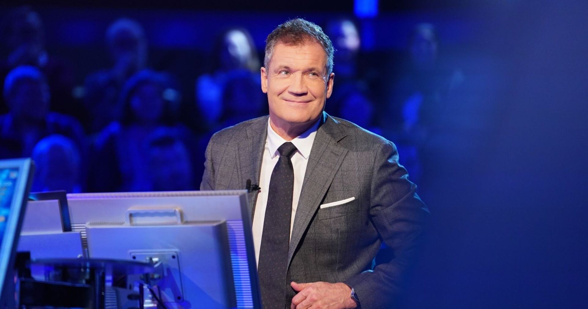Armin Assinger im Anzug bei der Moderation der „Millionenshow“, im Studio vor Publikum und Monitoren.