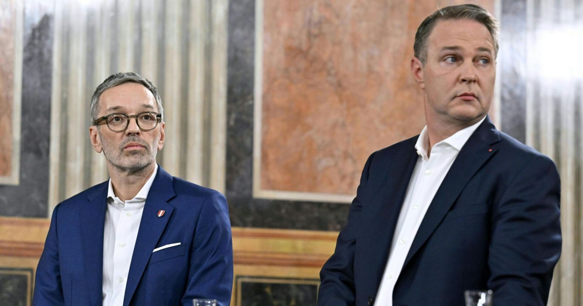 Herbert Kickl und Andreas Babler sitzen nebeneinander in einem prunkvollen Saal, beide in dunklen Anzügen, mit ernsten Mienen und Blicken in entgegengesetzte Richtungen.