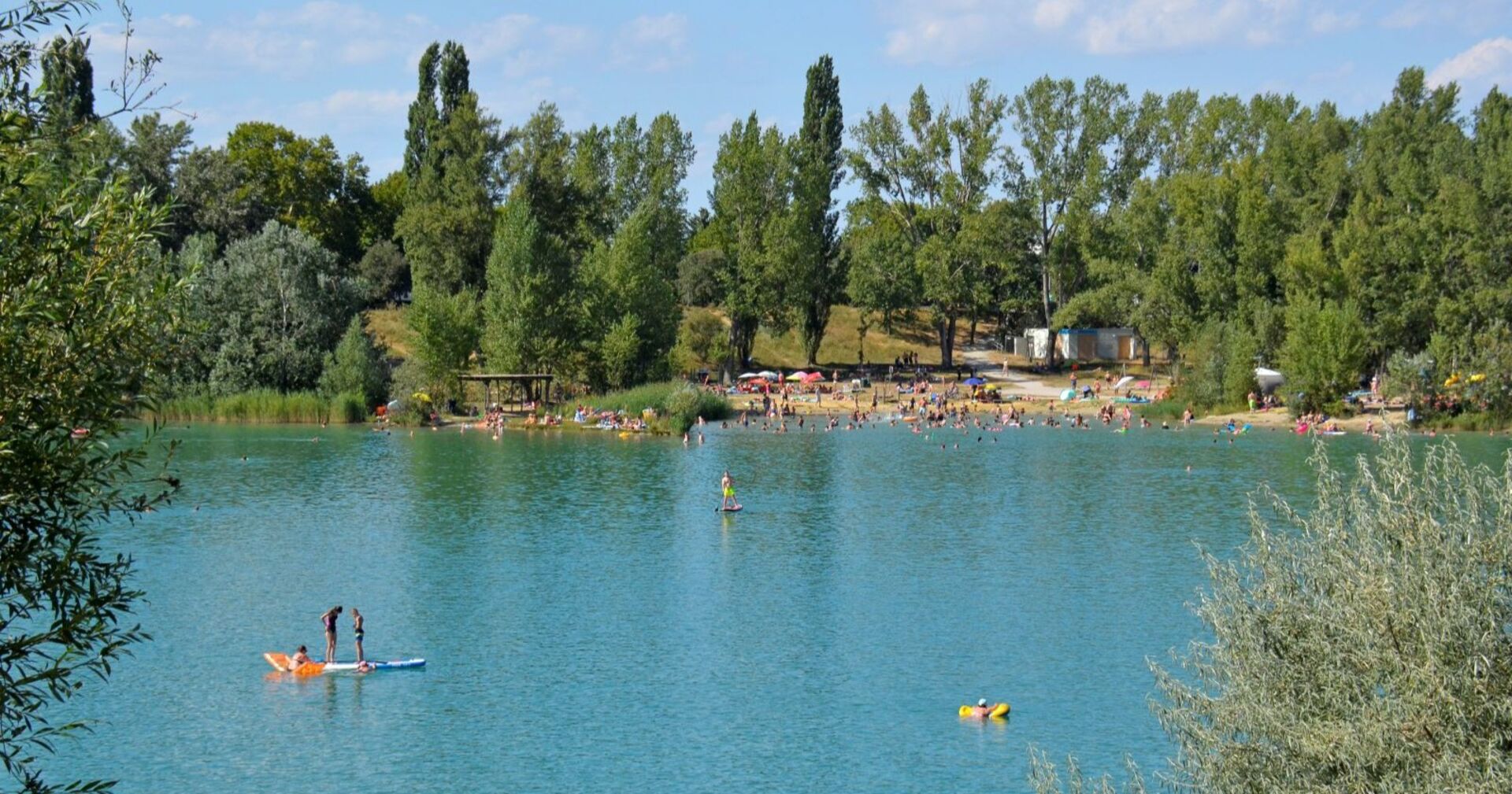 Badeteich Hirschstetten in Wien an einem sonnigen Tag, viele Menschen schwimmen und entspannen am Ufer, im Vordergrund Kinder auf einem Stand-Up-Paddle.