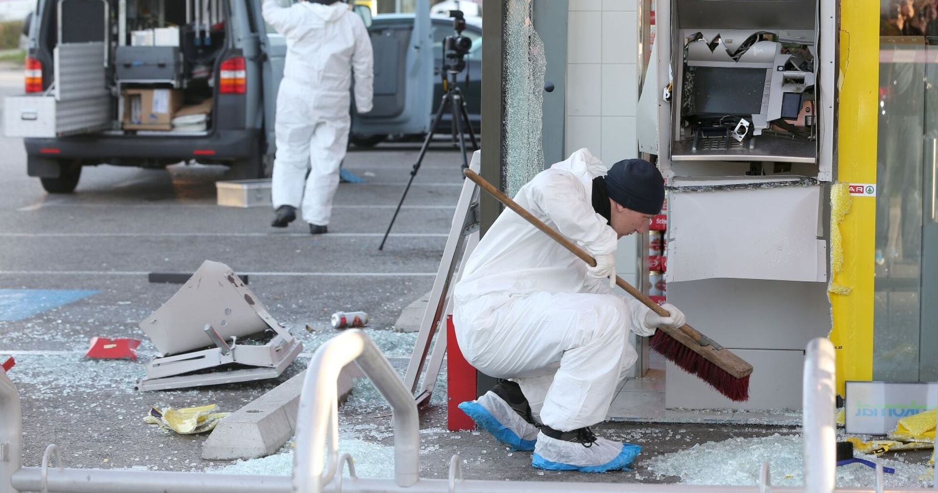 Ein Tatort nach einer Bankomat-Sprengung: Eine Person in weißem Spurensicherungsanzug fegt Glasscherben vor einem stark beschädigten Geldautomaten zusammen. Im Hintergrund sind weitere Ermittler sowie ein geöffnetes Einsatzfahrzeug mit Ausrüstung zu sehen.