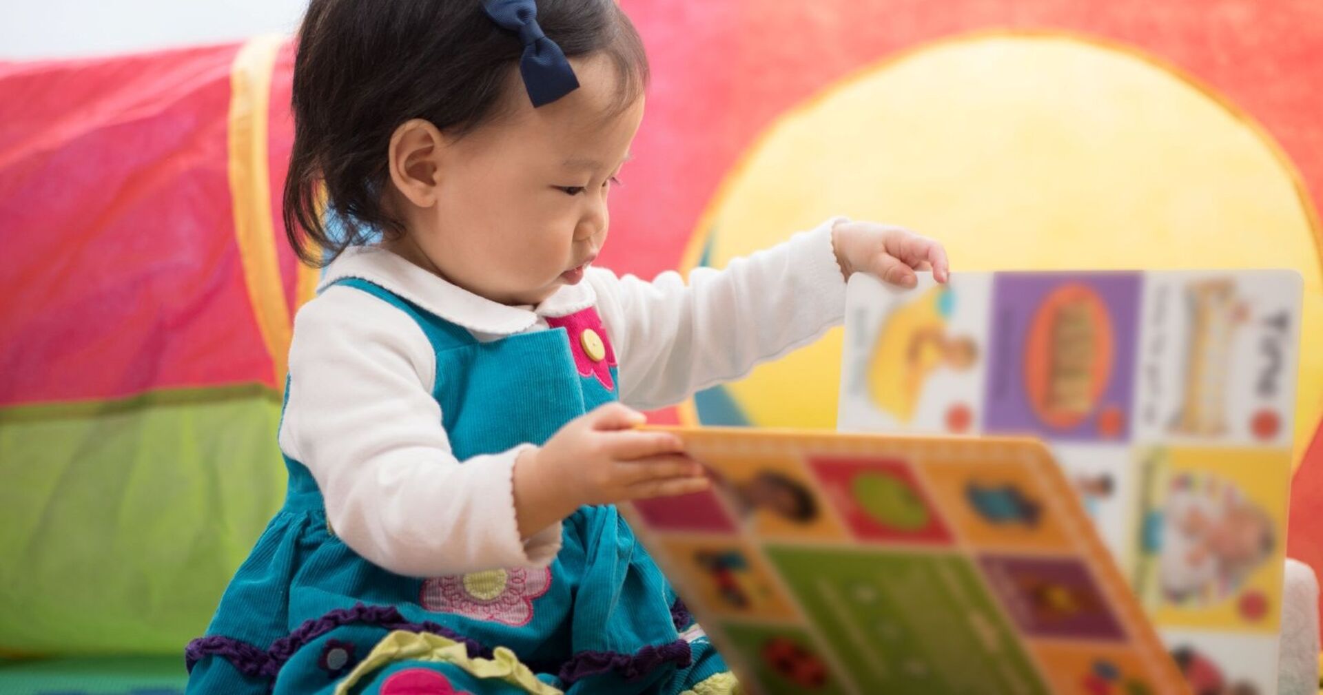 Kleines Mädchen mit dunklen Haaren und blauem Kleid sitzt in einem bunt dekorierten Raum und schaut in ein aufgeschlagenes Bilderbuch.