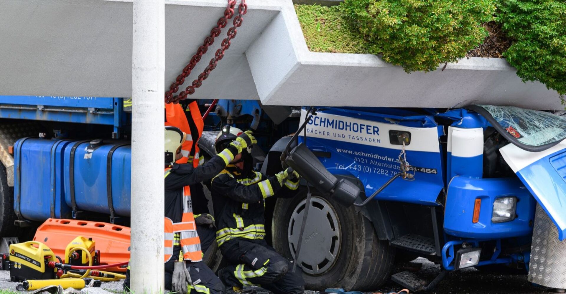 Zwei Feuerwehrleute arbeiten unter einem eingestürzten Brückenelement an einem stark beschädigten blauen Lkw. Die Fahrerkabine ist zerdrückt, Werkzeuge und Rettungsgeräte liegen am Boden.