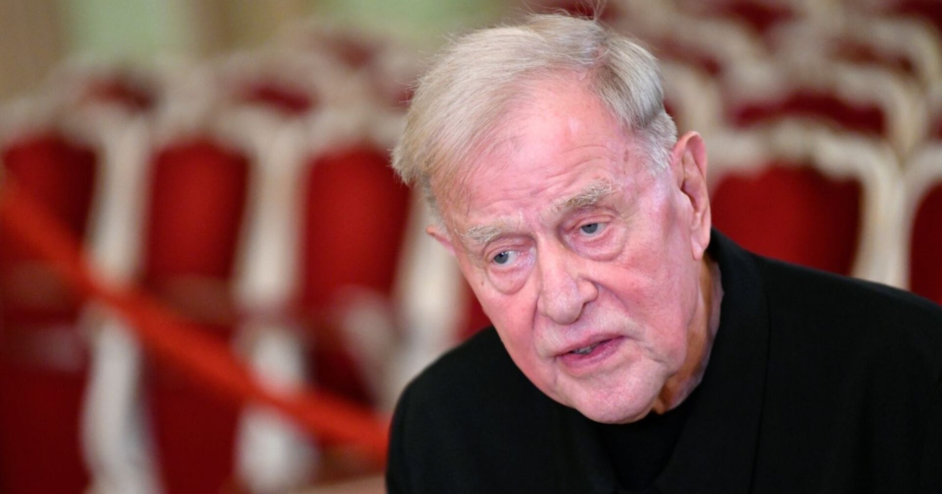 Der gealterte Claus Peymann mit ernstem Blick vor roten Theatersesseln – Großaufnahme des Regisseurs in einem Theatersaal.