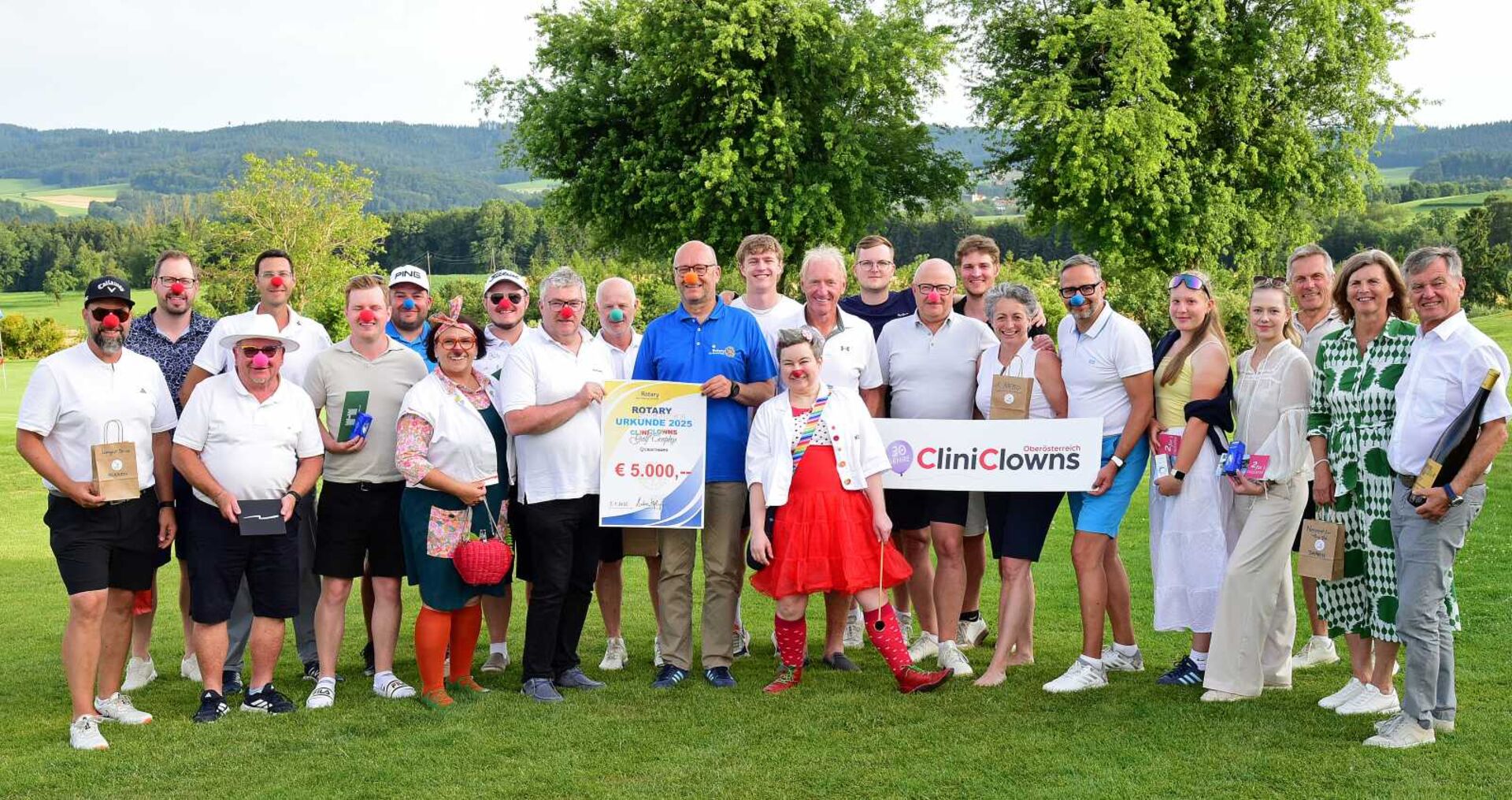 Gruppenfoto auf einem Golfplatz: Menschen mit roten Clownnasen halten einen 5.000 € Spendenscheck und ein CliniClowns-Schild.