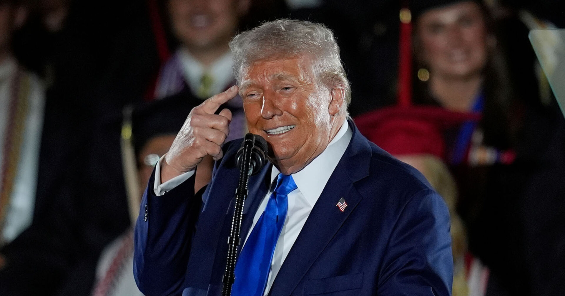 Donald Trump, im Anzug und mit Krawatte, hält sich Finger an den Kopf und zeigt &quot;den Vogel&quot;