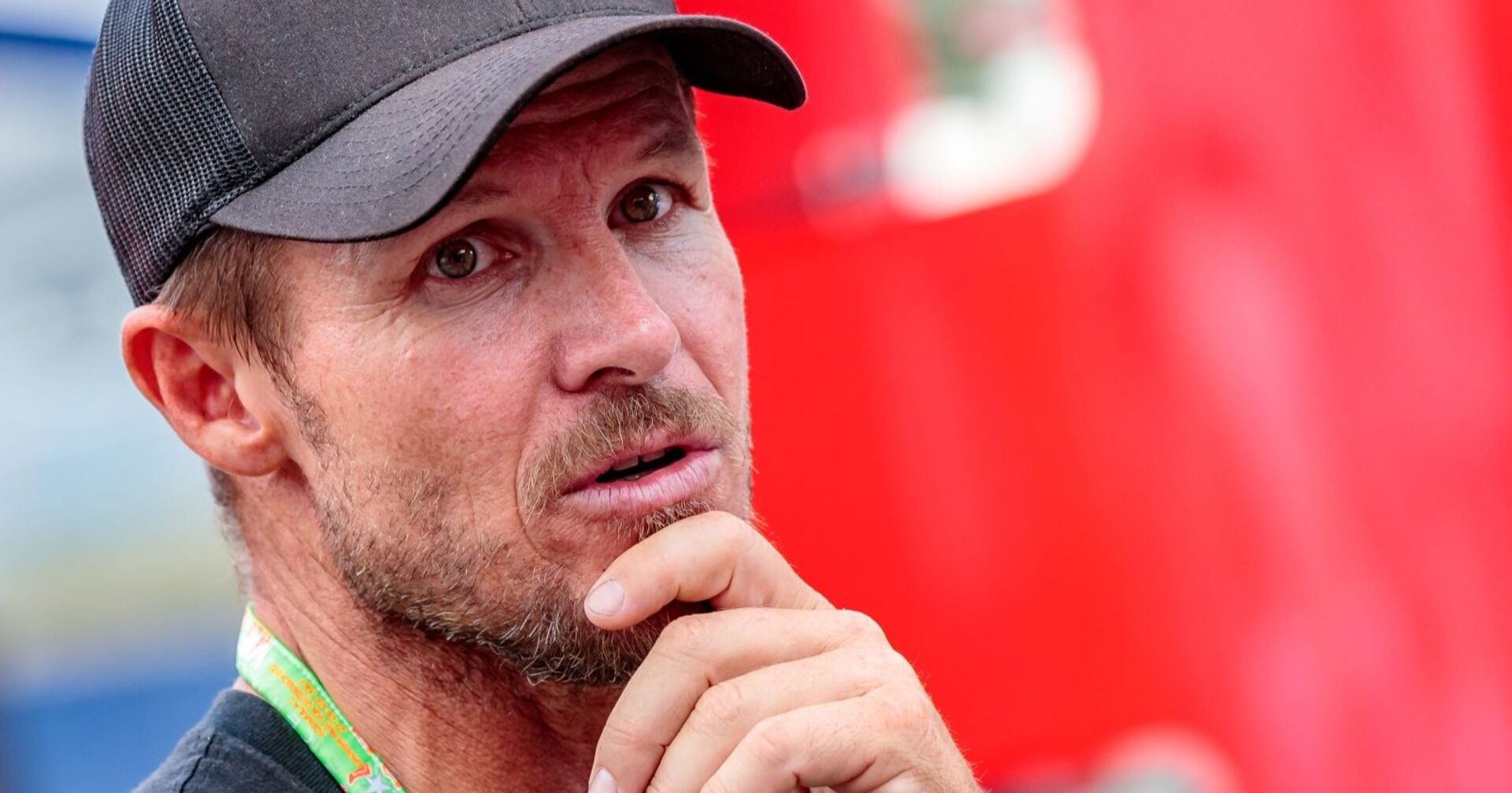 Porträt von Felix Baumgartner mit schwarzer Kappe, nachdenklicher Gesichtsausdruck, im Vordergrund seine Hand am Kinn, unscharfer roter Hintergrund.