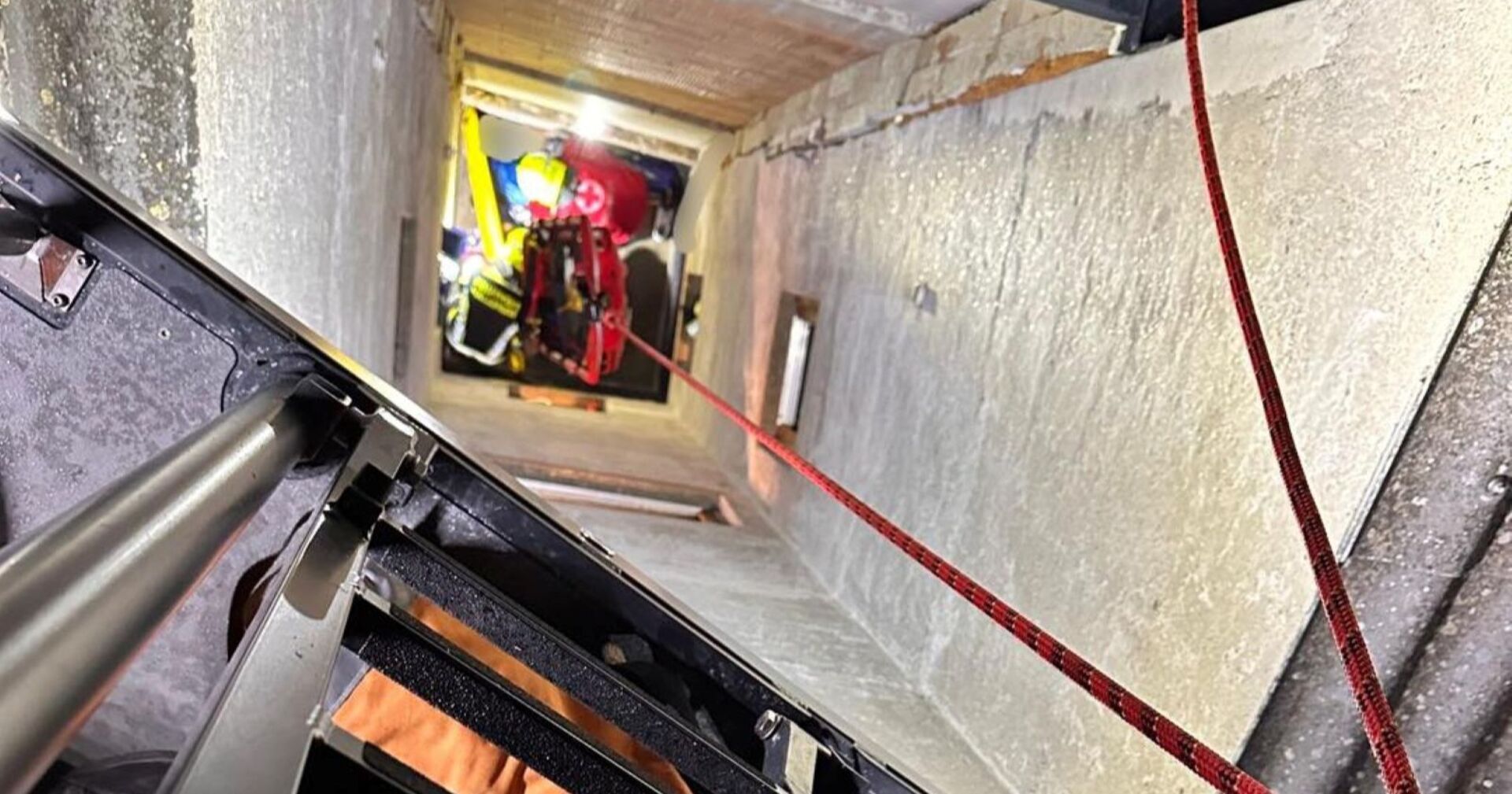 Blick in einen engen Schacht, in dem Feuerwehrleute mit einem roten Rettungskorb einen Jugendlichen nach oben ziehen. Ein rotes Sicherungsseil ist gespannt, eine Leiter ist im Vordergrund zu sehen.