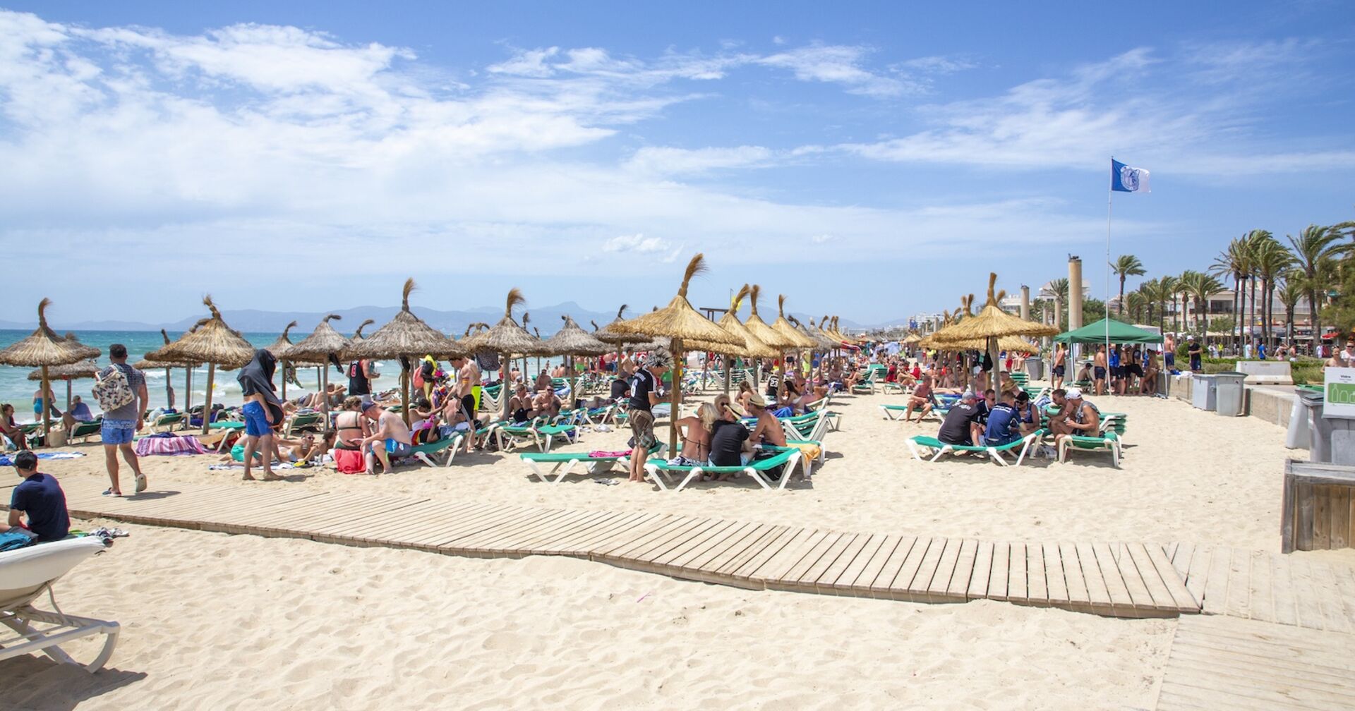 Menschen liegen und sitzen unter Strohdach-Sonnenschirmen an einem gut besuchten Strand auf Mallorca. Der Sand ist hell, das Meer türkisblau, eine blaue Flagge weht im Wind.