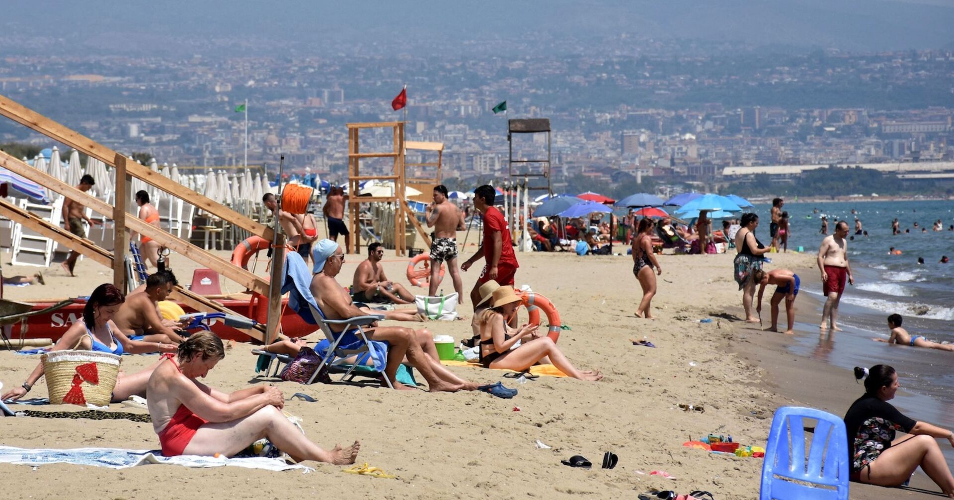 Menschen an einem überfüllten Sandstrand in Catania, Sizilien, bei extremer Sommerhitze; viele sitzen unter Sonnenschirmen oder gehen ins Meer zur Abkühlung.