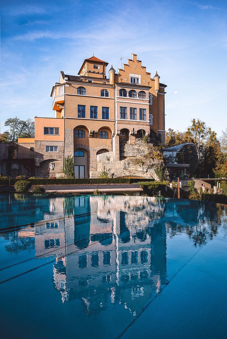 Fassade von Hotel Schloss Mönchstein in Salzburg, gespiegelt im Wasser des Infinity-Pools bei klarem Himmel.