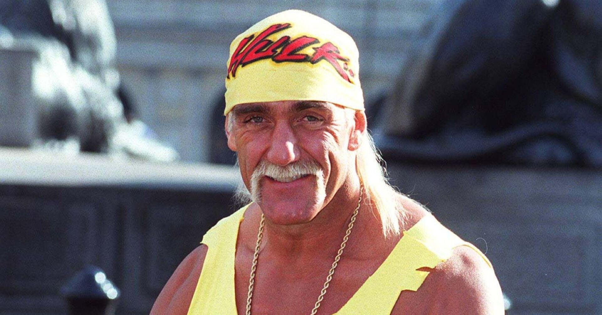 Hulk Hogan trägt ein gelbes Bandana mit roter Aufschrift und ein gelbes Muskelshirt. Er lächelt in die Kamera.