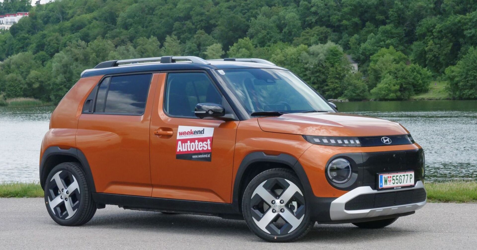 Ein kaffeebrauner Hyundai Inster parkt am Ufer eines Flusses. Am anderen Ufer ist ein dichter Laubwald zu sehen.