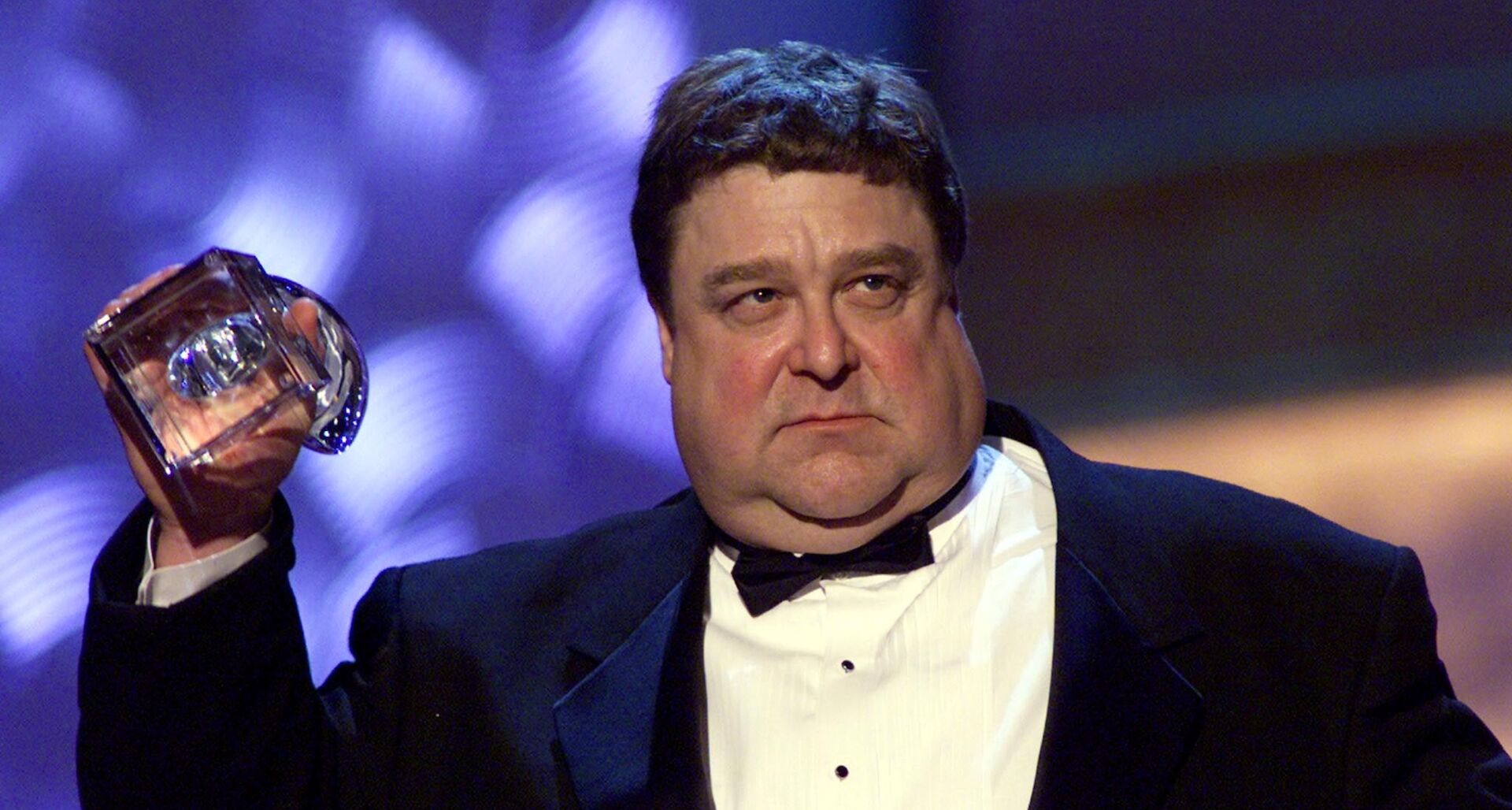 John Goodman früher stark übergewichtig im Smoking, mit ernstem Blick und Pokal in der Hand.