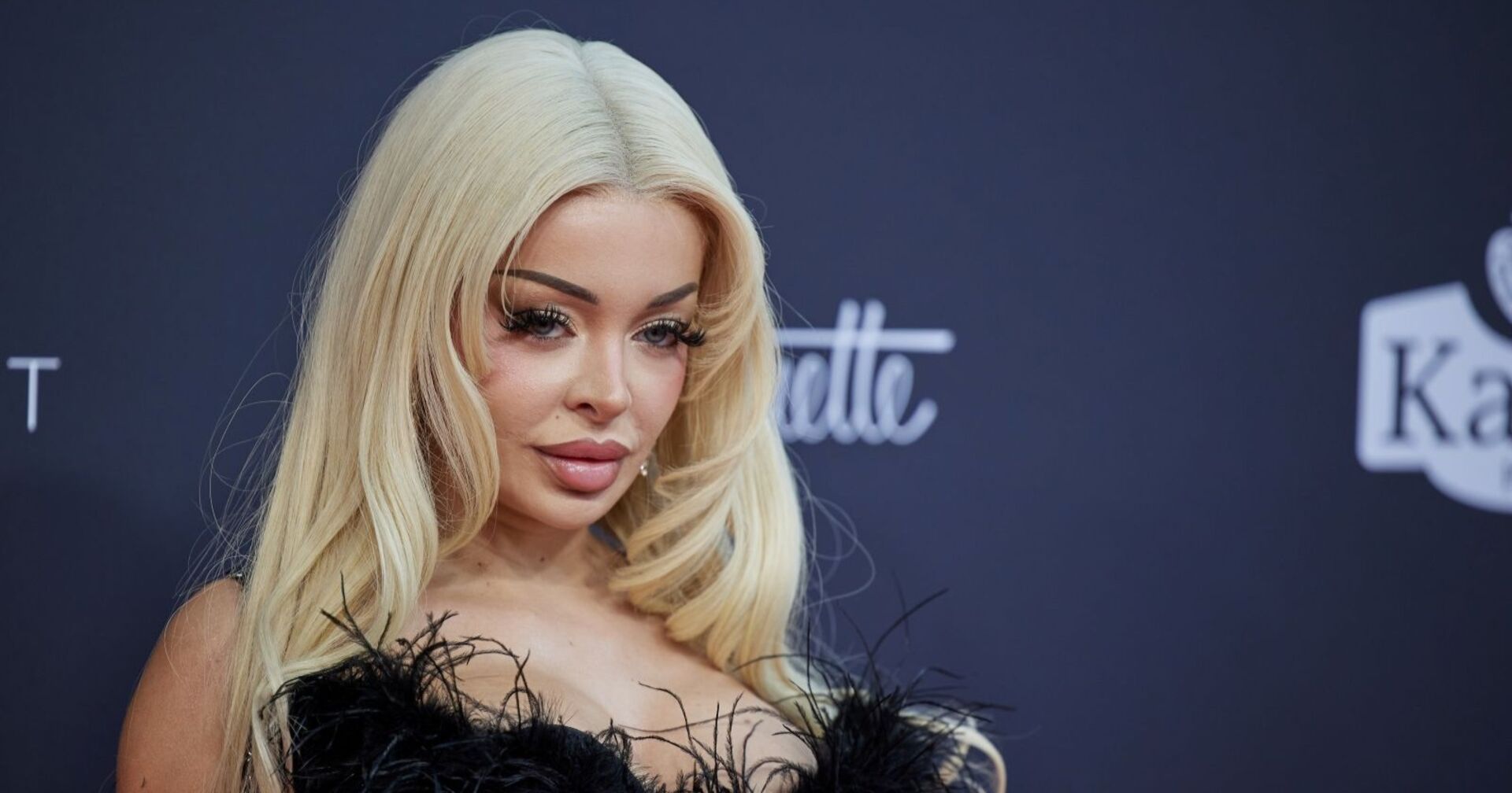 Katja Krasavice mit blondem Haar und tiefem Ausschnitt auf dem roten Teppich, schaut lasziv in die Kamera.