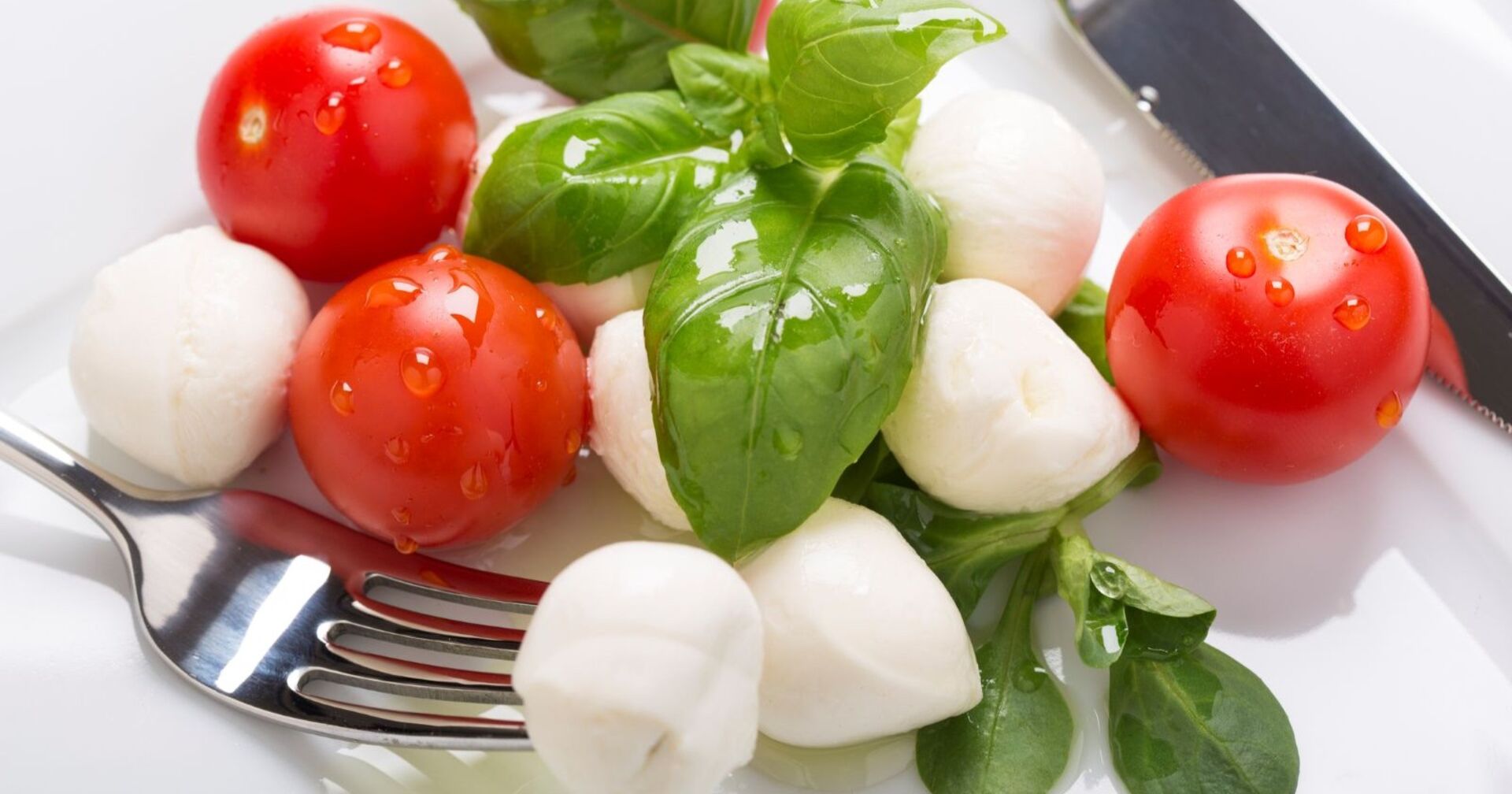 Nahaufnahme eines Tellers mit kleinen Mozzarellakugeln, frischen Cocktailtomaten, grünem Basilikum und einem Metallbesteck – eine typische Caprese-Vorspeise.