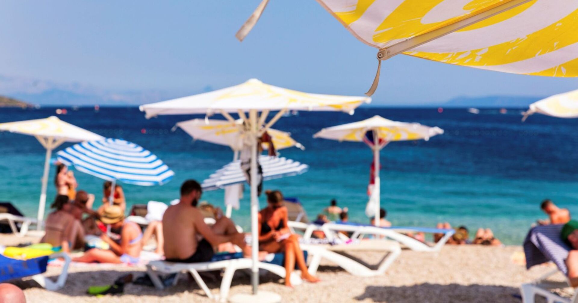 Aufnahme eines mediterranen Strandes mit unscharf zu erkennenden Badegästen, im Vordergrund ein Sonnenschirm