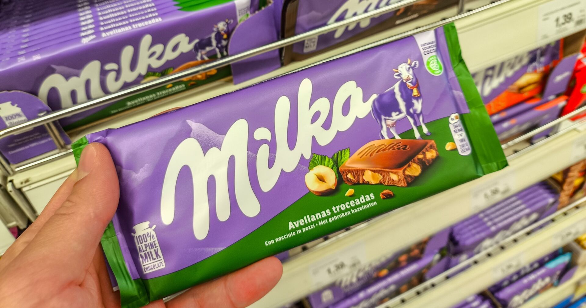 Hand hält eine Milka-Schokoladentafel mit Haselnüssen im Supermarkt, dahinter weitere Milka-Tafeln in lila Verpackung im Regal.