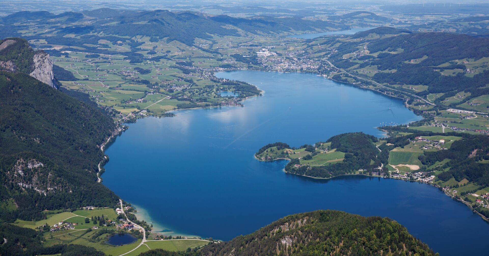 Luftaufnahme des Mondsees in Oberösterreich, umgeben von grünen Hügeln und Dörfern. Deutlich zu sehen sind die Uferbereiche mit Stegen, Buchten und angrenzender Infrastruktur.