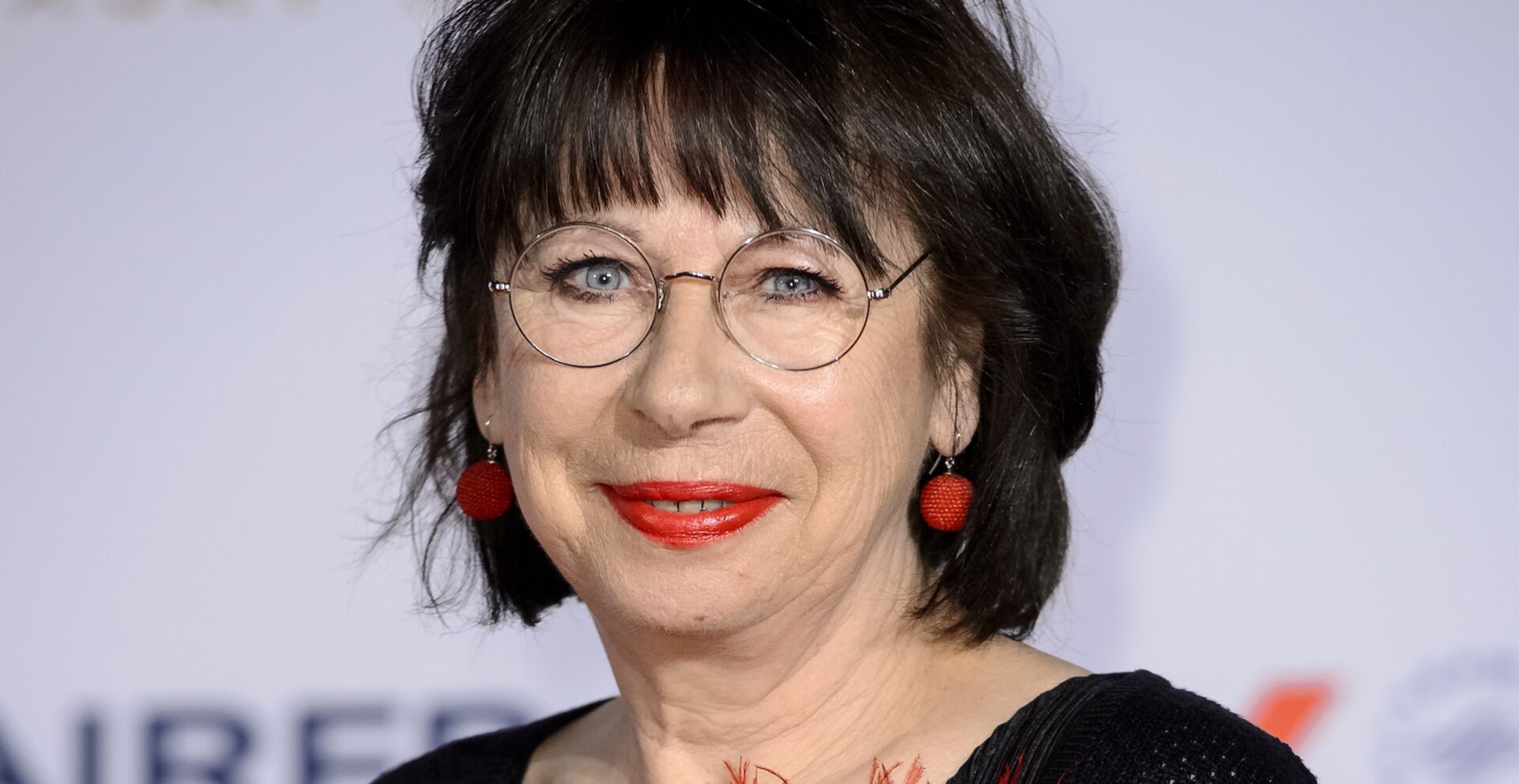 Porträt von Monika Hansen mit schwarzem Haar, Brille und rotem Lippenstift, lächelnd bei einem Auftritt in Berlin.