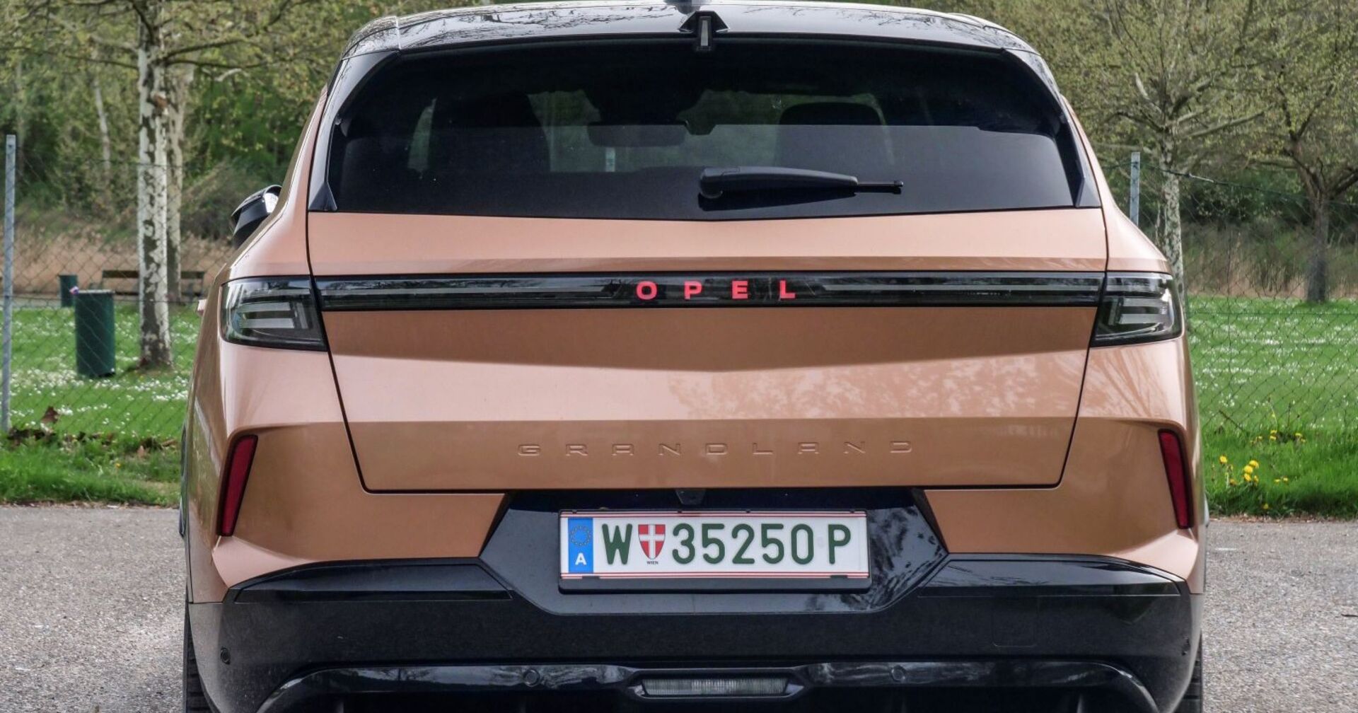 Das Heck des Opel Grandland, dessen Farbe an Milchschokolade erinnert. Das Auto steht vor einer Wiese, die mit einem Maschendrahtzaun eingezäunt ist. Auf der Wiese stehen einige Obstbäume.
