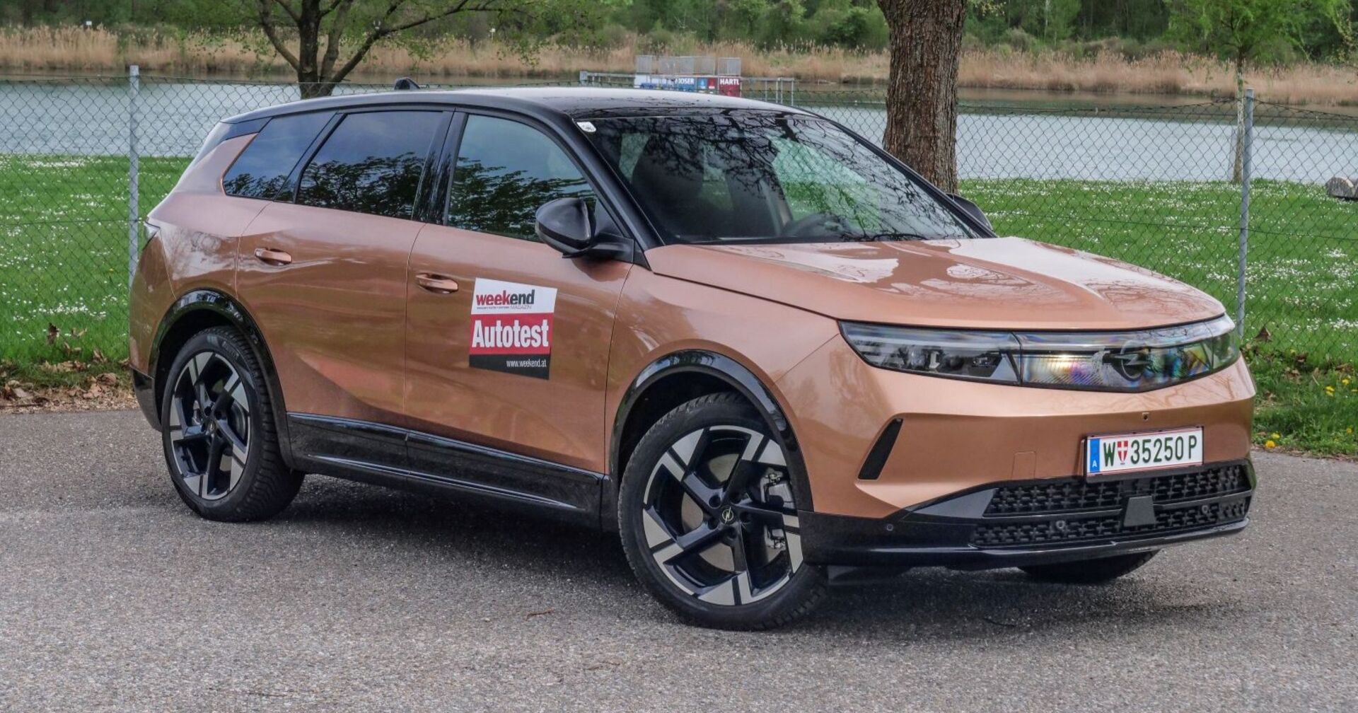 Ein Opel Grandland, dessen Farbe an Milchschokolade erinnert, steht auf einem Parkplatz. Im Hintergrund sind eine Wiese und ein Gewässer zu erkennen.