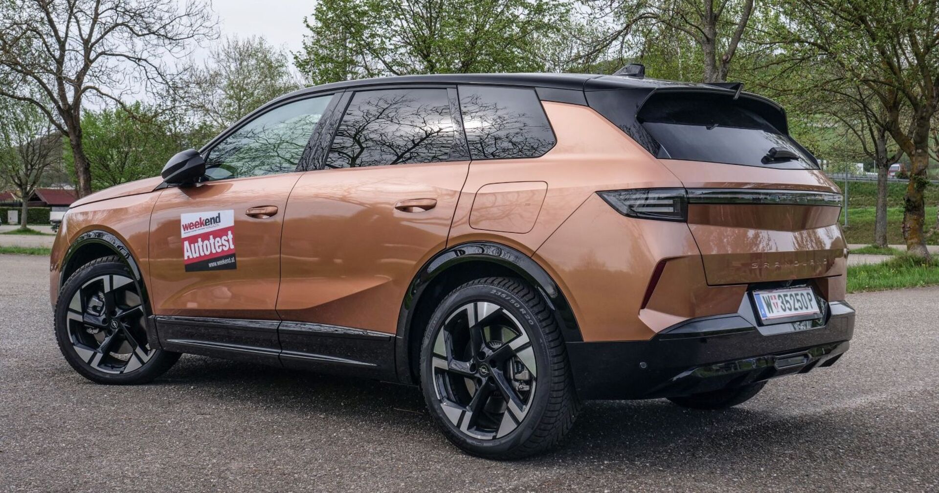 Der Opel Grandland, dessen Farbe an Milchschokolade erinnert, von der Seite. Das Auto steht auf einem asphaltierten  Parkplatz, im Hintergrund sind einige Laubbäume zu sehen, die auf einer Wiese stehen.
