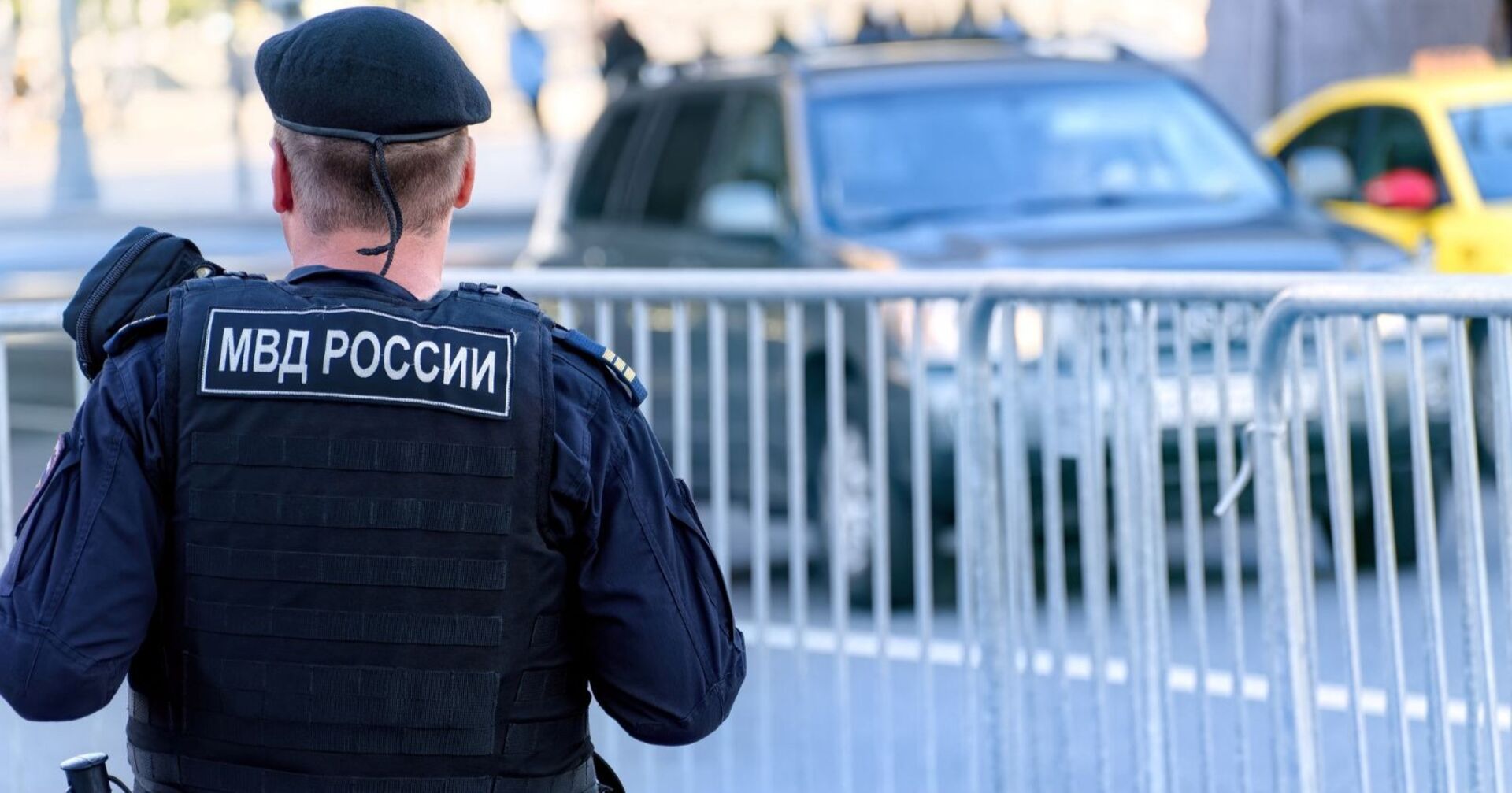Russischer Polizist der Nationalgarde in kugelsicherer Weste patrouilliert durch die Straßen der Stadt, vor ihm ist ein Zaun, dahinter ein Auto