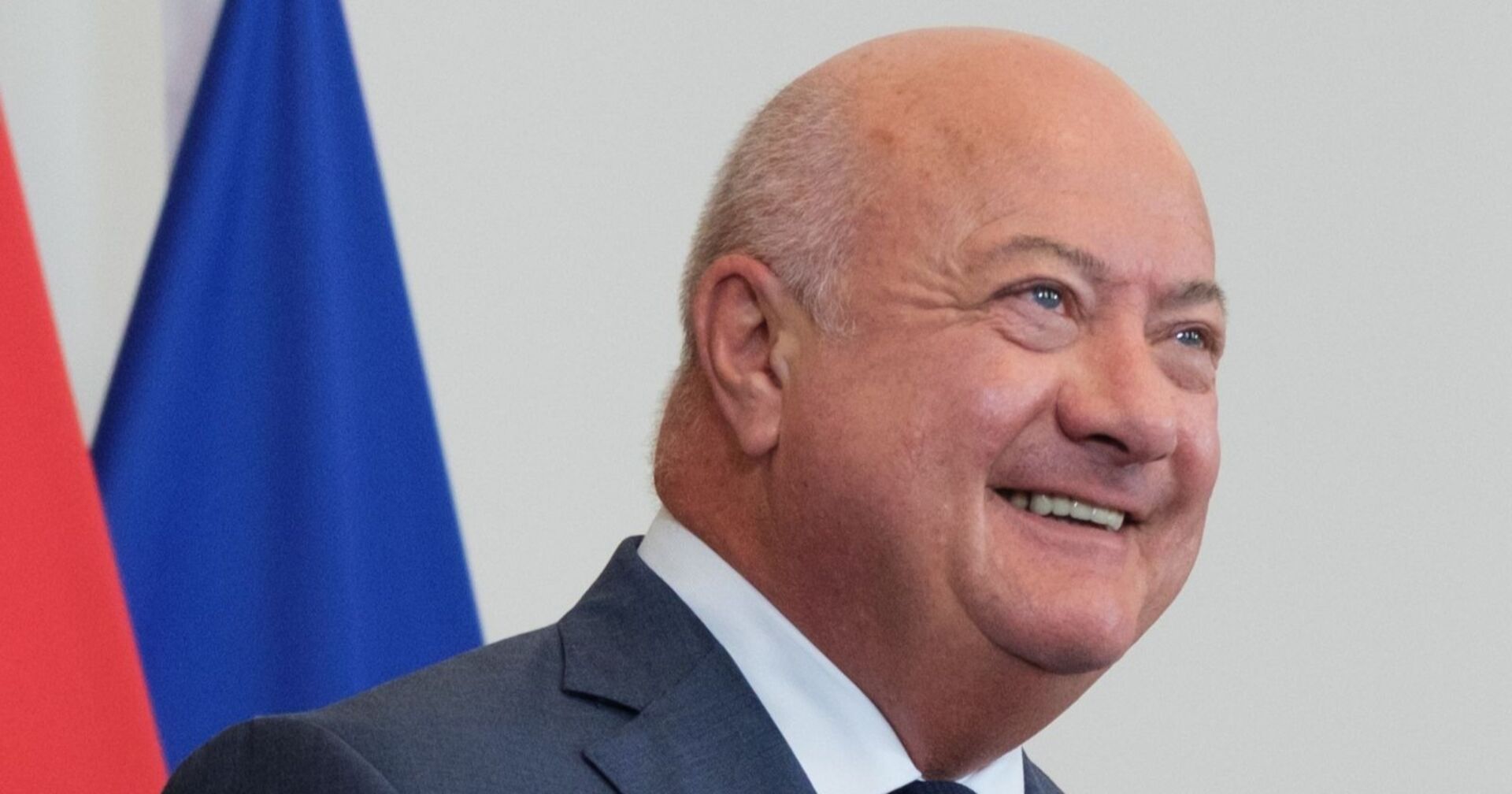 Porträt von Kanzler Christian Stocker, lachend im Anzug, im Hintergrund unscharf rote und blaue Flaggen.