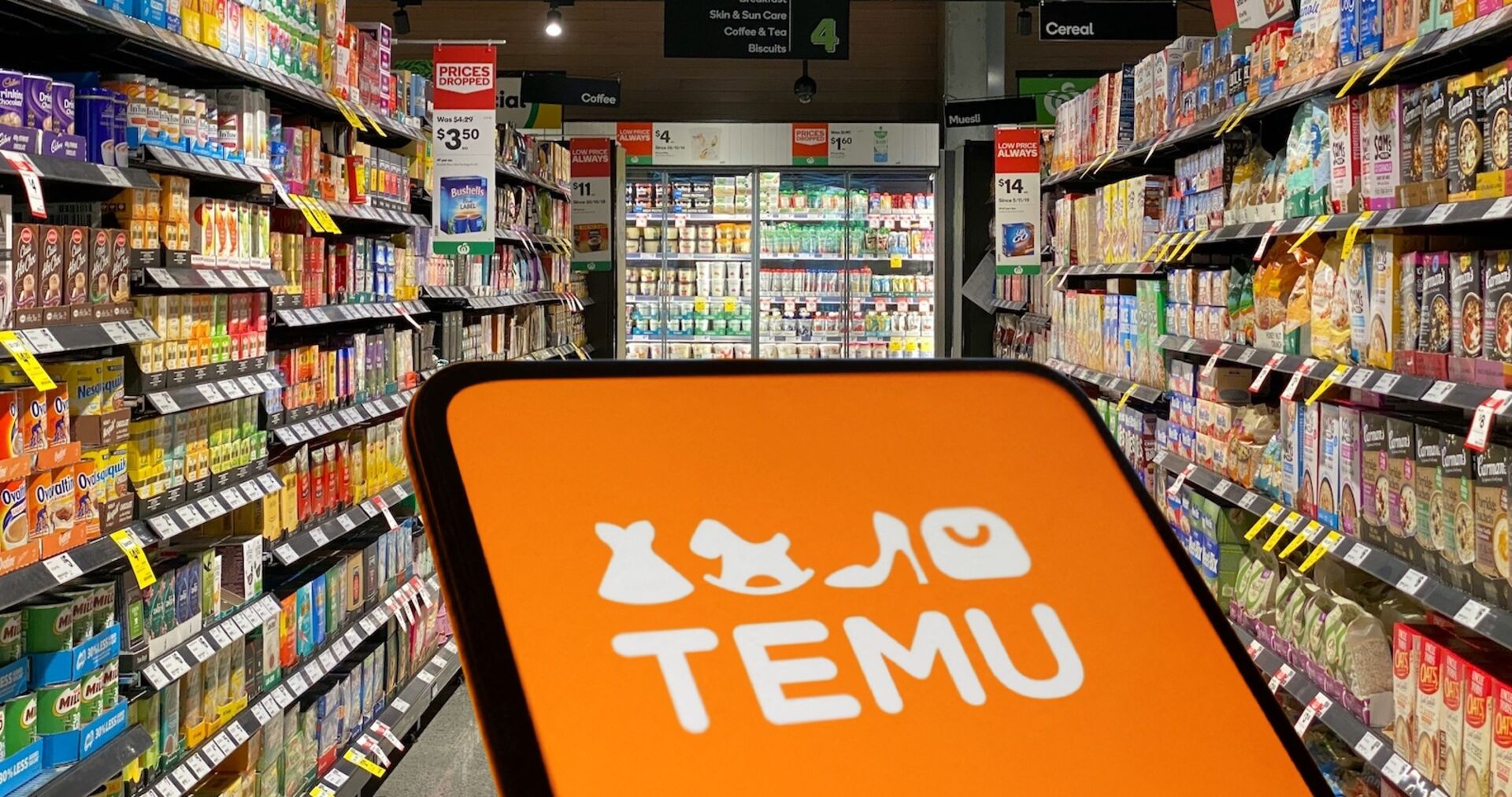 Ein Smartphone mit dem orangefarbenen Temu-Logo im Vordergrund, dahinter ein Supermarktgang mit Regalen voller Lebensmittelpackungen und Kühlregalen.