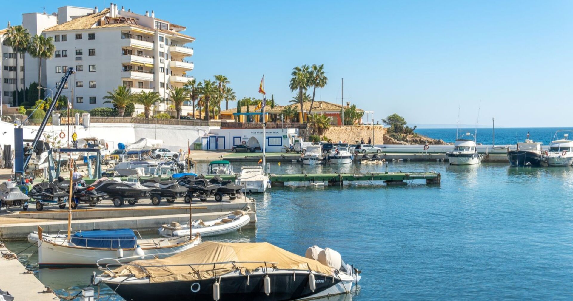 Blick auf einen kleinen Yachthafen in Palma Nova, Mallorca, mit Booten im Wasser, Palmen, einem Hotelgebäude und blauem Himmel im Hintergrund.