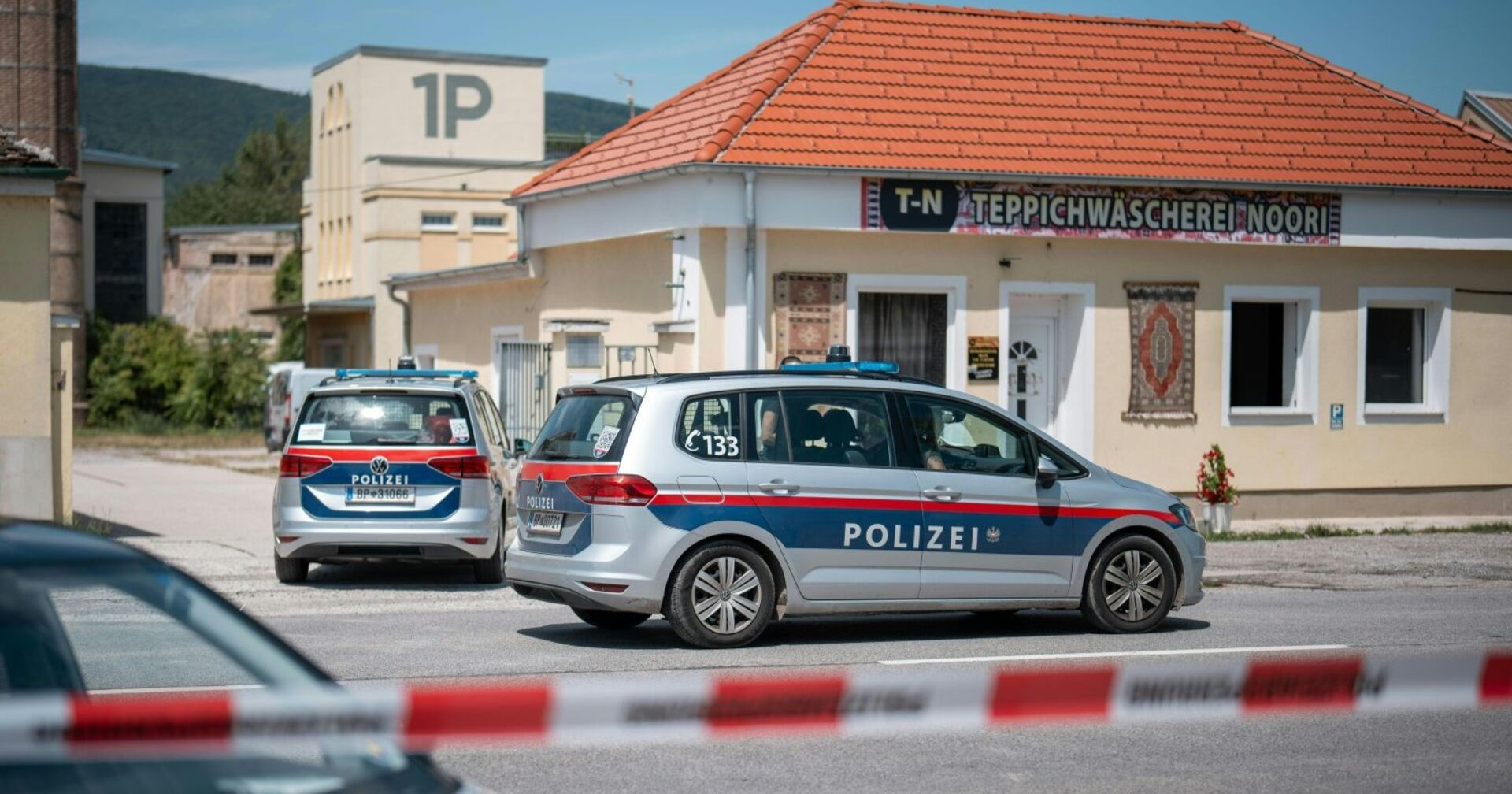 Zwei österreichische Polizeiautos stehen vor einem Gebäude in Traiskirchen mit der Aufschrift „Teppichwäscherei Noori“. Der Bereich ist mit rot-weißem Absperrband gesichert. Im Hintergrund sind Industriegebäude und grüne Hügel zu sehen.