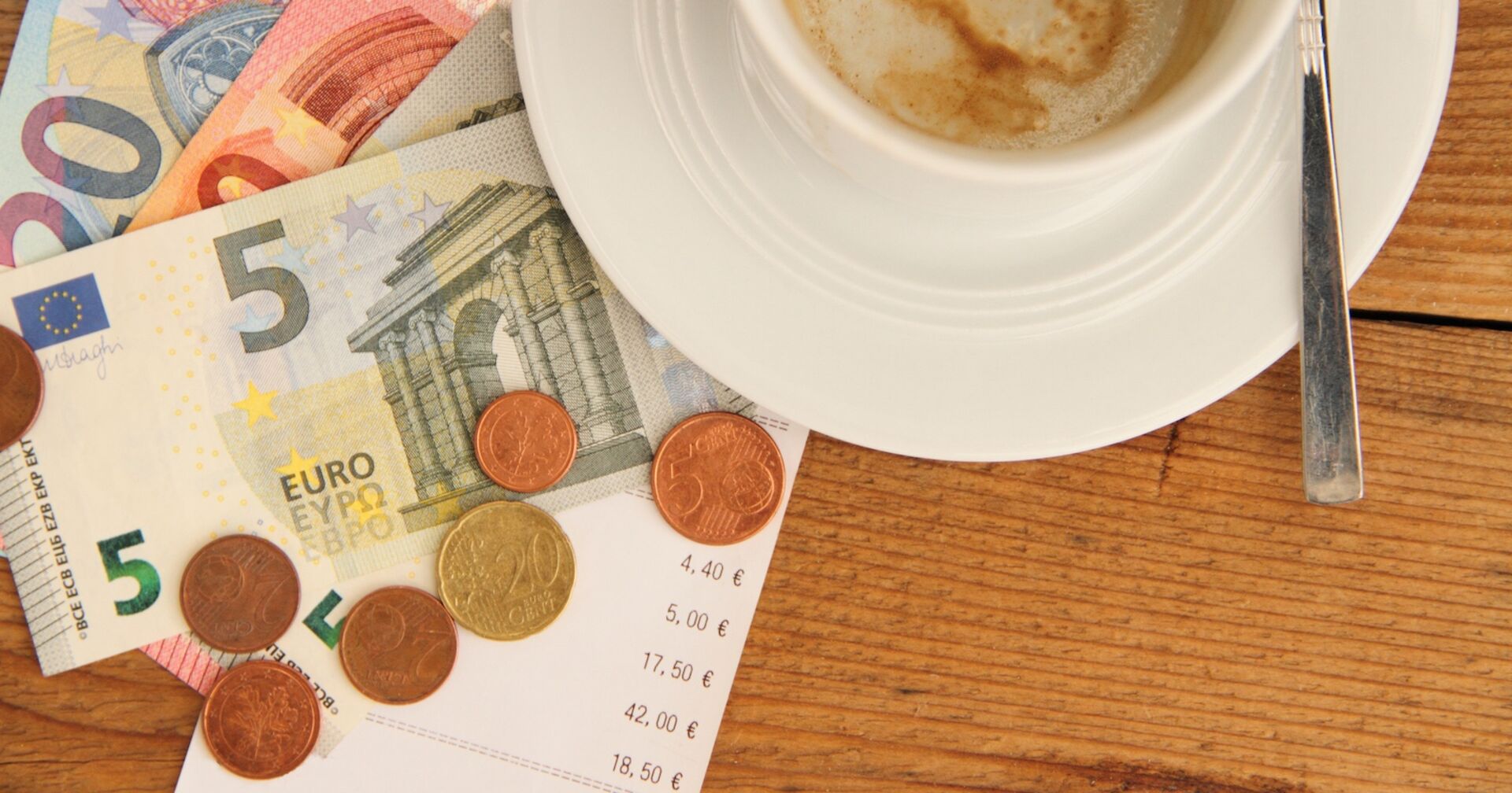 Mehrere Euro-Scheine und Münzen liegen auf einer Restaurantrechnung neben einer weißen Kaffeetasse mit Löffel auf einem Holztisch – Symbolbild für Trinkgeld in der Gastronomie.