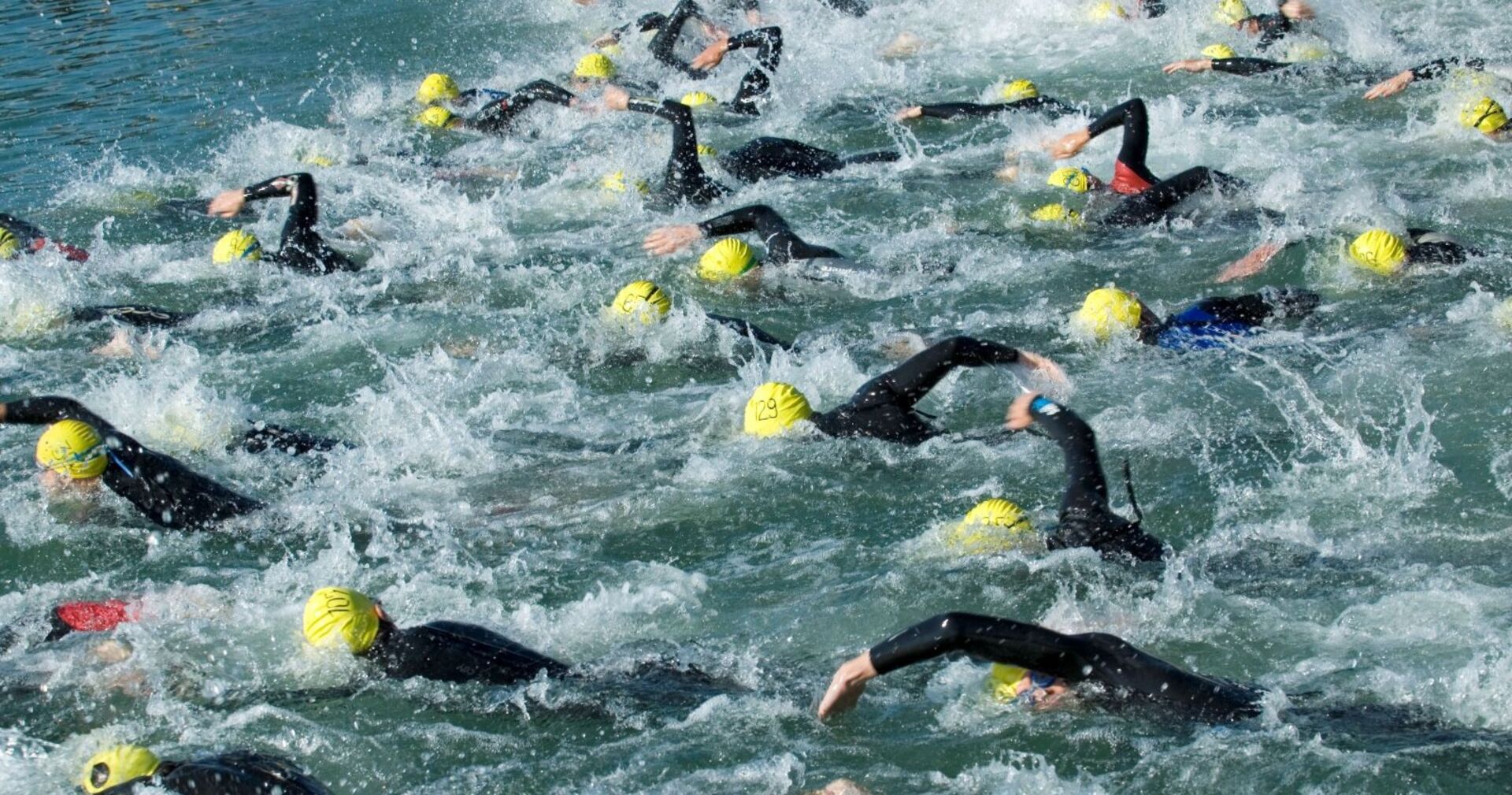 Zahlreiche Triathlet:innen mit gelben Badekappen und schwarzen Neoprenanzügen schwimmen eng nebeneinander im offenen Wasser eines Sees. Die Szene zeigt starke Wasserbewegung und Wettkampf-Stimmung.