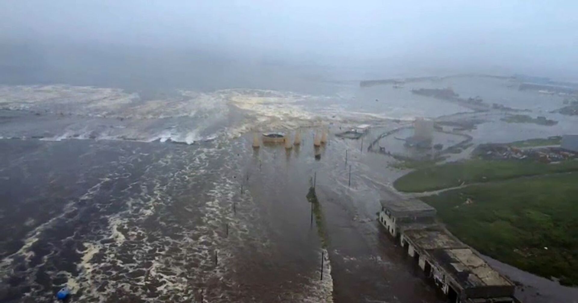 Luftaufnahme einer überfluteten Küstenstadt bei starkem Wellengang: Eine breite Tsunami-Welle überschwemmt Straßen, Häuser und Hafenanlagen unter grauem Himmel.
