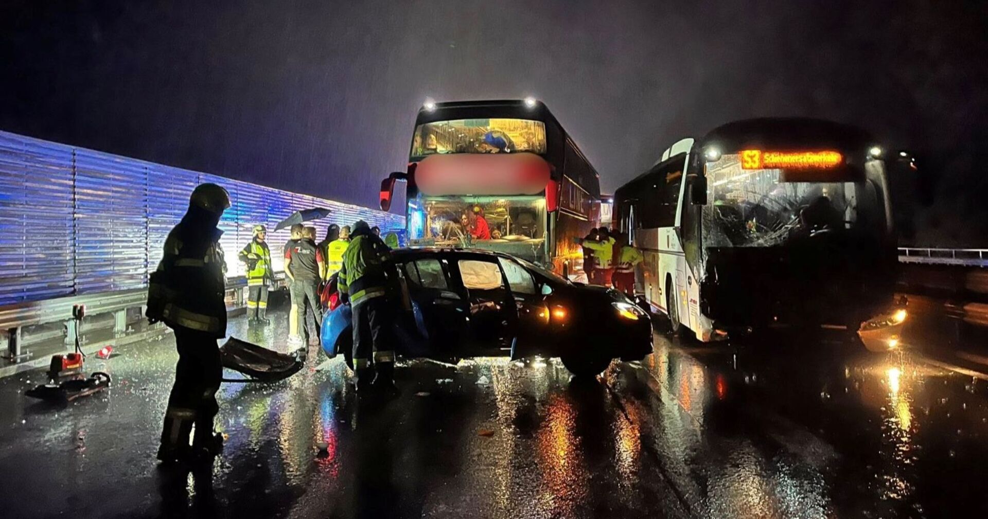 Unfallstelle bei Nacht auf der A10 Tauernautobahn: Zwei Busse und ein stark beschädigter Pkw stehen nach einem Crash auf regennasser Straße, Feuerwehrleute und Einsatzkräfte sichern die Unfallstelle.