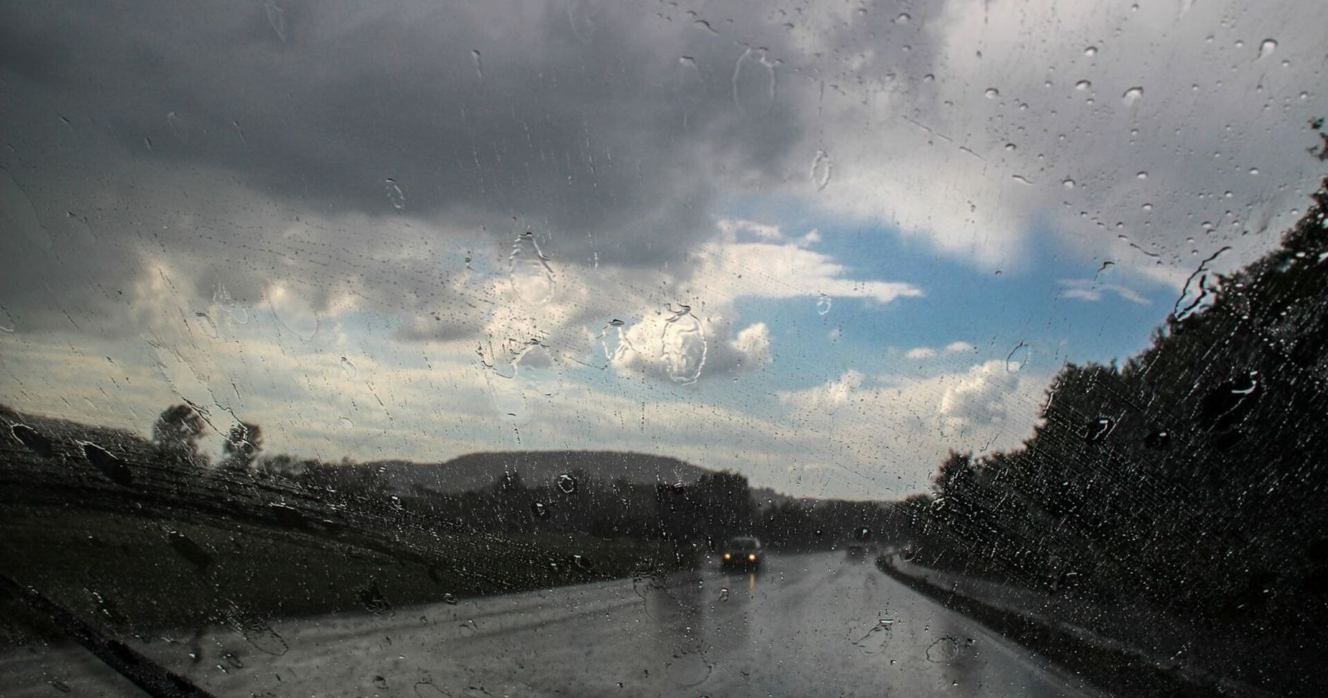 Blick durch eine regennasse Windschutzscheibe auf eine Landstraße, dunkle Gewitterwolken ziehen auf, dazwischen zeigen sich noch blaue Himmelsflecken.