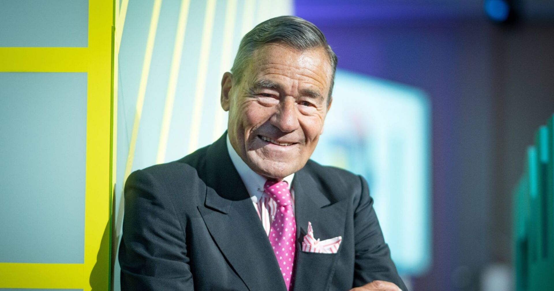 Wolfgang Grupp, elegant gekleidet im dunklen Anzug mit pinker Krawatte, steht lächelnd mit verschränkten Armen vor einer modernen Kulisse.