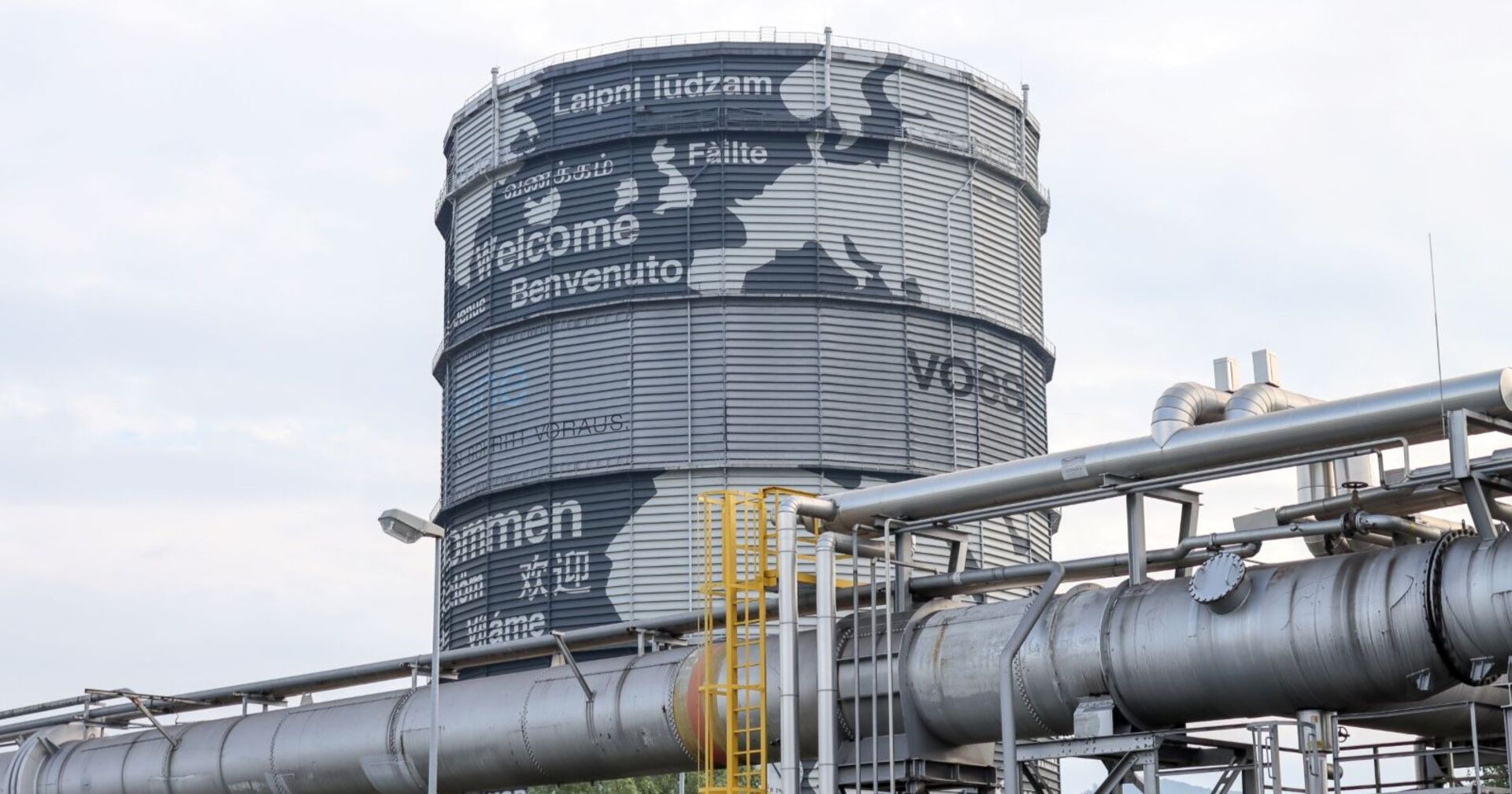 Großer, runder Gasspeicher bei der voestalpine in Linz mit internationalen Willkommensgrüßen, umgeben von industriellen Rohrleitungen.