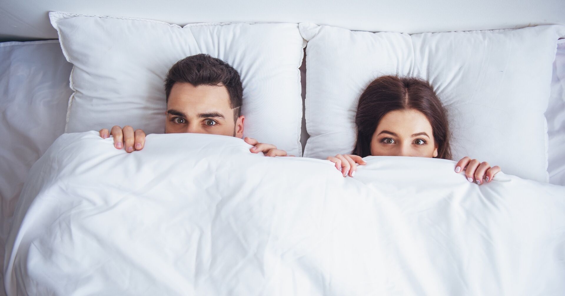 Ein junges Paar liegt im Bett und zieht sich die Decke bis zur Nase.