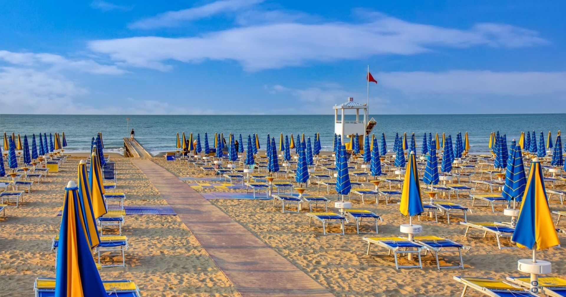 Leerer Strandabschnitt in Jesolo mit blauen und gelben Sonnenschirmen, Blick auf das Meer und einen Rettungsturm unter roter Flagge