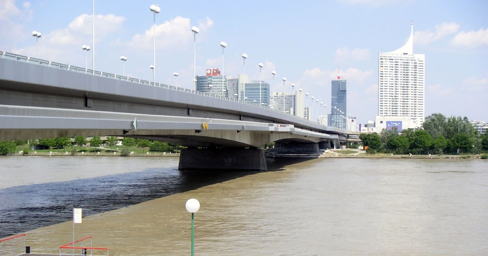 Blick auf die Reichsbrücke in Wien über der Donau, im Hintergrund moderne Hochhäuser der Donau City, vorne Wasser mit sichtbarer Strömungsgrenze.